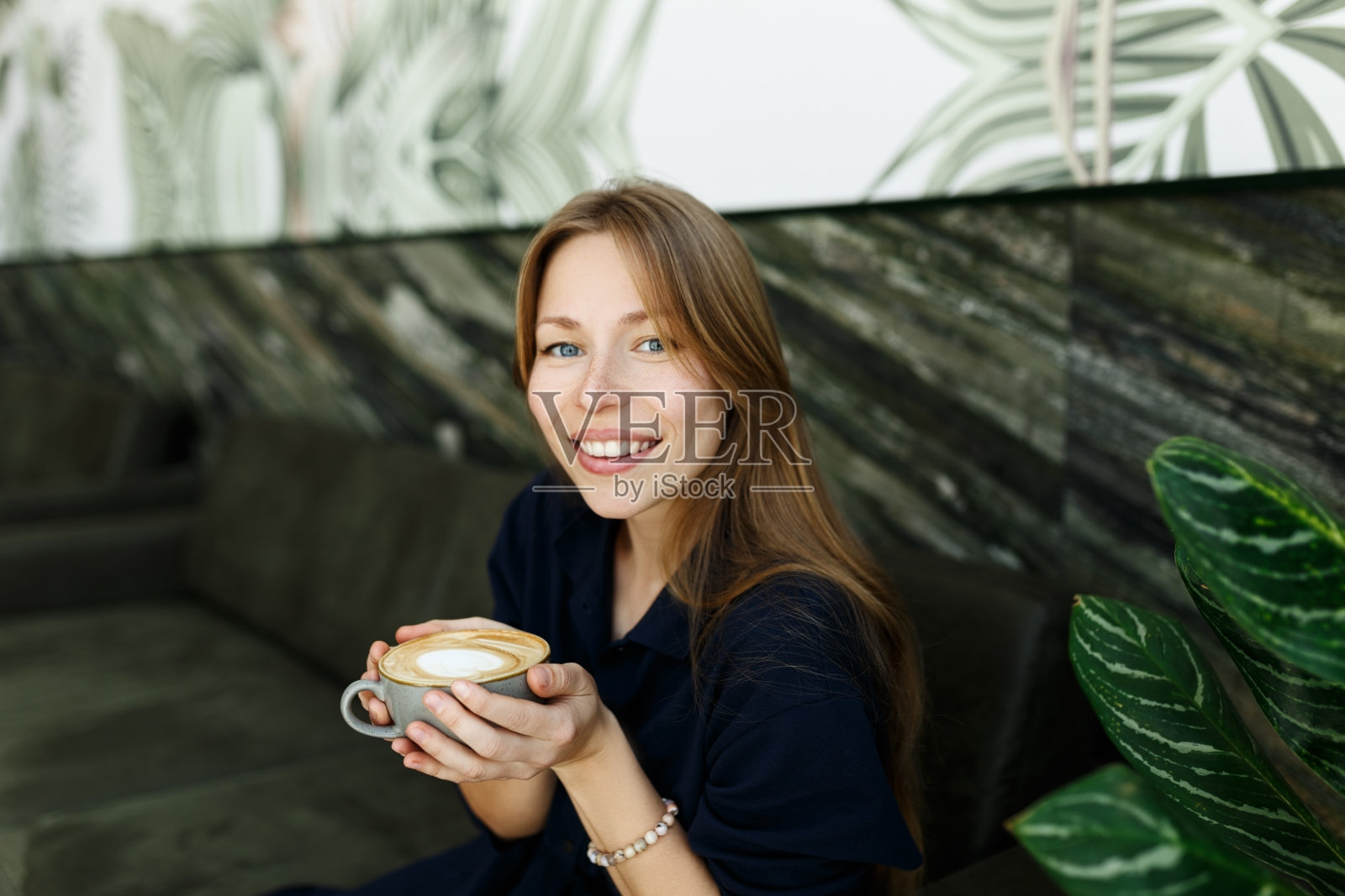 快乐的年轻女子在家里喝咖啡的画像。微笑的漂亮女孩喝着热饮。兴奋的女人穿着绿色内饰的蓝裙子，闻着卡布奇诺的味道照片摄影图片