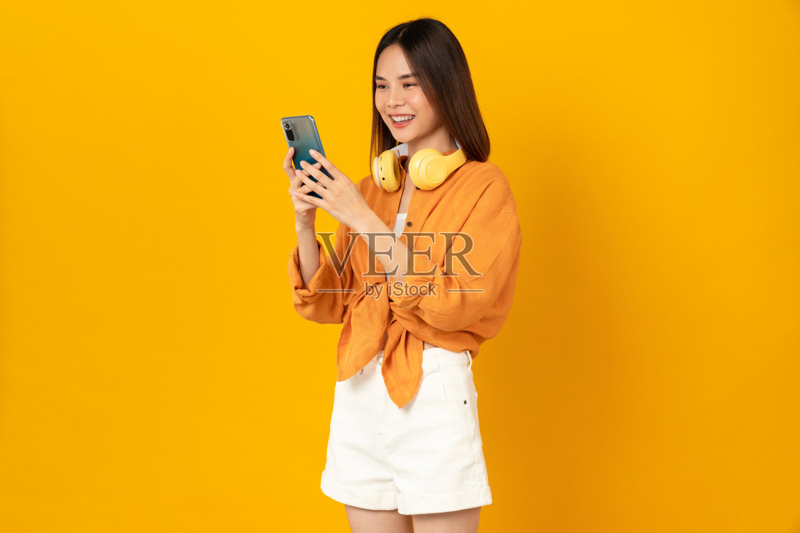 欢快的年轻亚洲女子戴着耳机听音乐，享受智能手机上最喜欢的播放列表应用程序，在橙色背景下跳舞。照片摄影图片