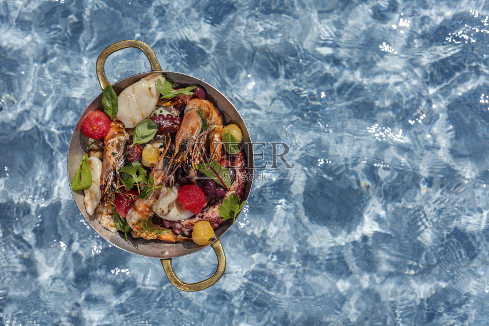 各式各样的海鲜、卡巴马尔、章鱼、鲈鱼、虾、烤西红柿和香草、西红柿、橄榄和橄榄油一起放在青铜模具里。盘子放在水中央的架子上。照片摄影图片