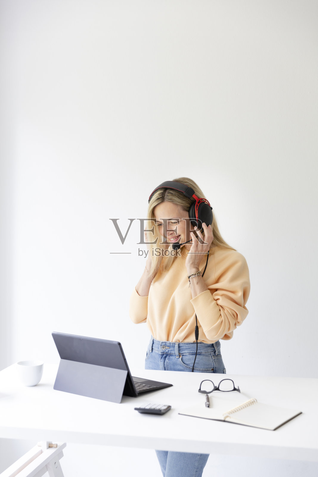 漂亮的金发女郎头戴耳机站在高桌旁，穿着高腰裤，通过笔记本电脑与客户交谈照片摄影图片