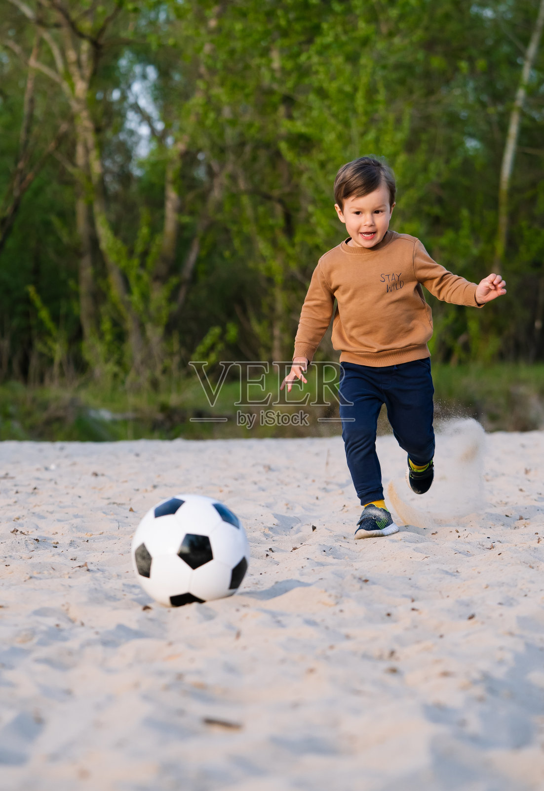 有趣的小男孩在沙滩上的公园里玩黑白足球。小孩跑上去用脚踢球照片摄影图片