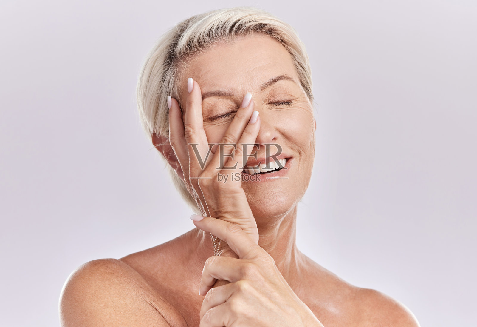 一个快乐的成熟白人女性放松的手在她的脸对粉红色的背景。微笑的年长女性休息，照顾她的皮肤，同时做一个护肤程序照片摄影图片