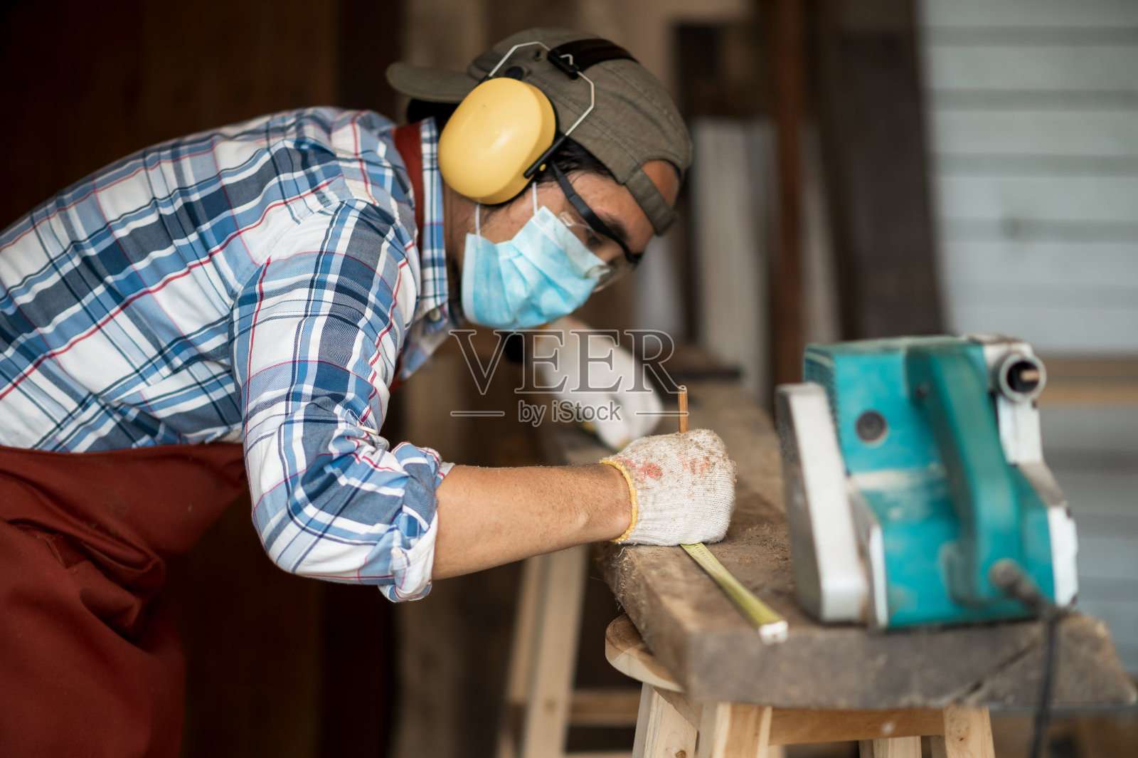 一位年轻的亚洲木匠，系着棕色的围裙，在木板上工作时，使用磨砂机和卷尺。木匠概念在木材工厂工作。有选择性的重点。照片摄影图片