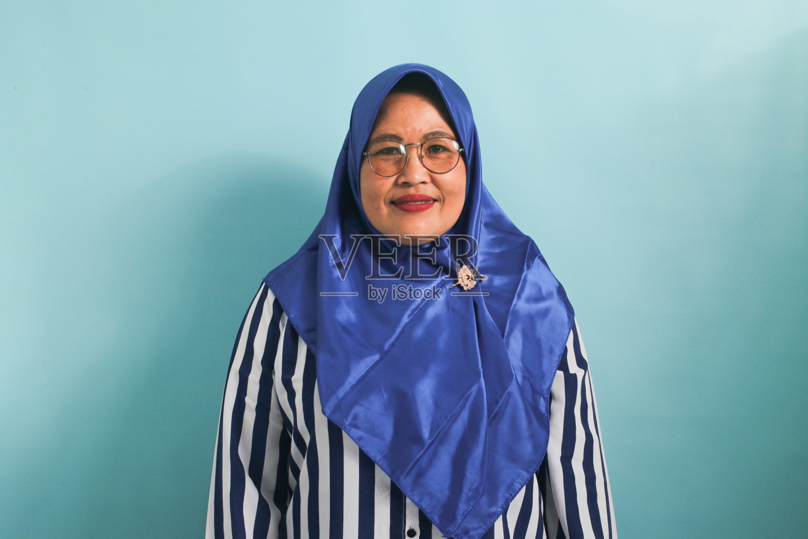 戴着头巾，戴着眼镜的中年亚洲妇女，在蓝色的背景下孤独地看着镜头。照片摄影图片