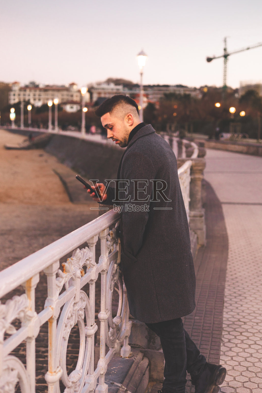 日落时分，在多诺斯塔-圣塞巴斯蒂安海滨散步，一名年轻的白人男子在使用智能手机。照片摄影图片