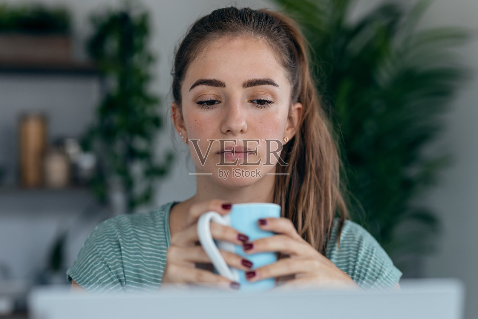 漂亮的年轻女子在家里的厨房边喝咖啡边用笔记本电脑工作。照片摄影图片