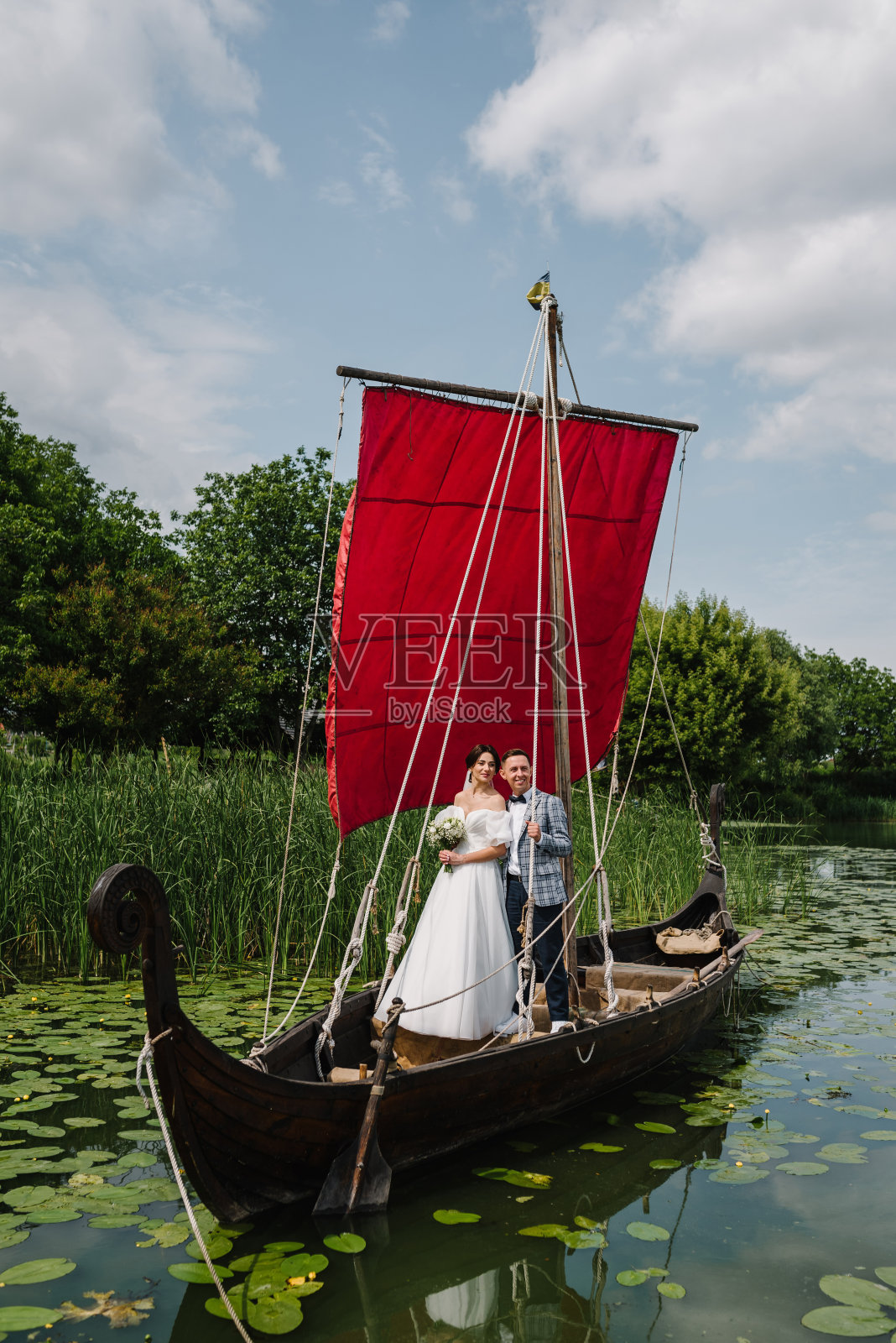 年轻的新婚夫妇在海盗船上拥抱并摆姿势。新娘和新郎在质朴的小船上。新婚快乐。在户外结婚。在湖边或池塘旁举行婚礼。照片摄影图片
