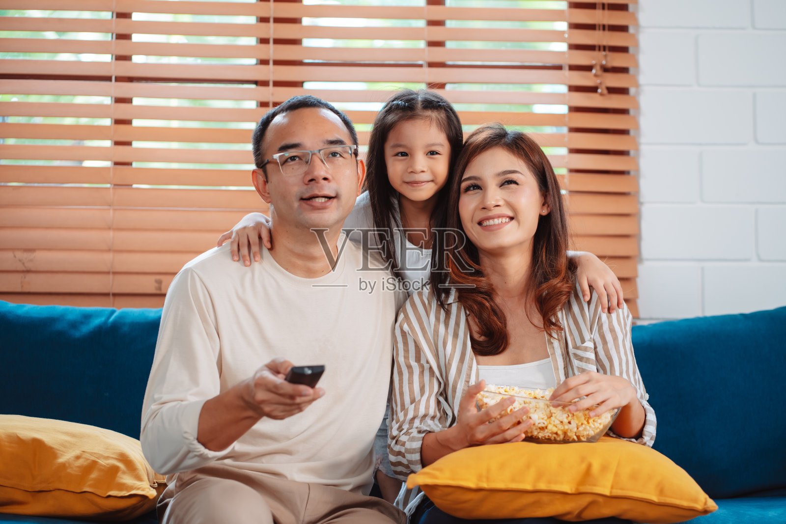 亚洲家庭由父母和女儿在家里客厅的沙发上看电视或电影，吃爆米花组成。享受放松的快乐。照片摄影图片