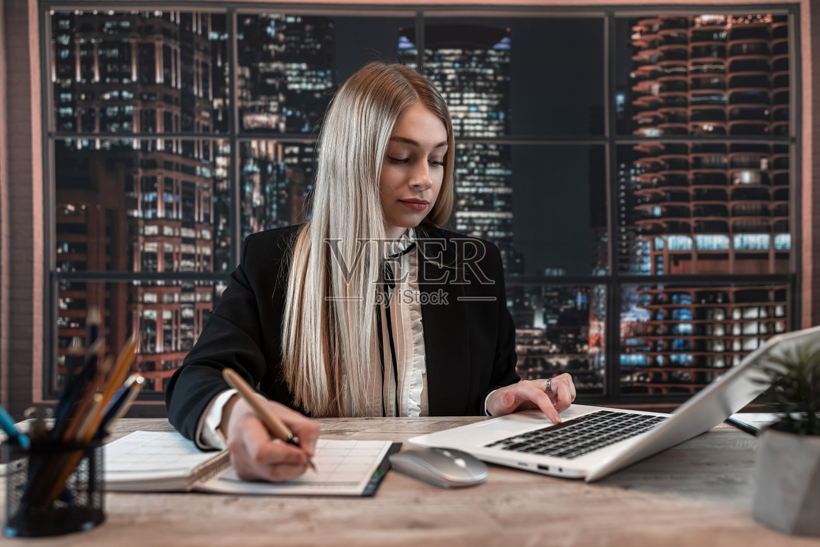 年轻女子穿着黑色西装坐在桌边，在办公室里拿着空的记事本和笔记本电脑工作。照片摄影图片