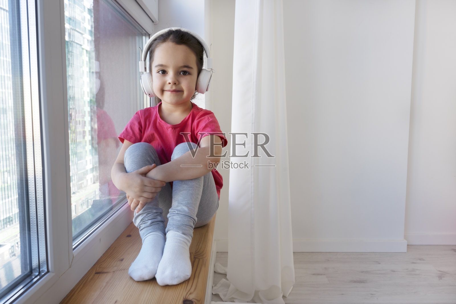 可爱快乐的小女孩戴着无线耳机坐在新公寓的窗台上。照片摄影图片