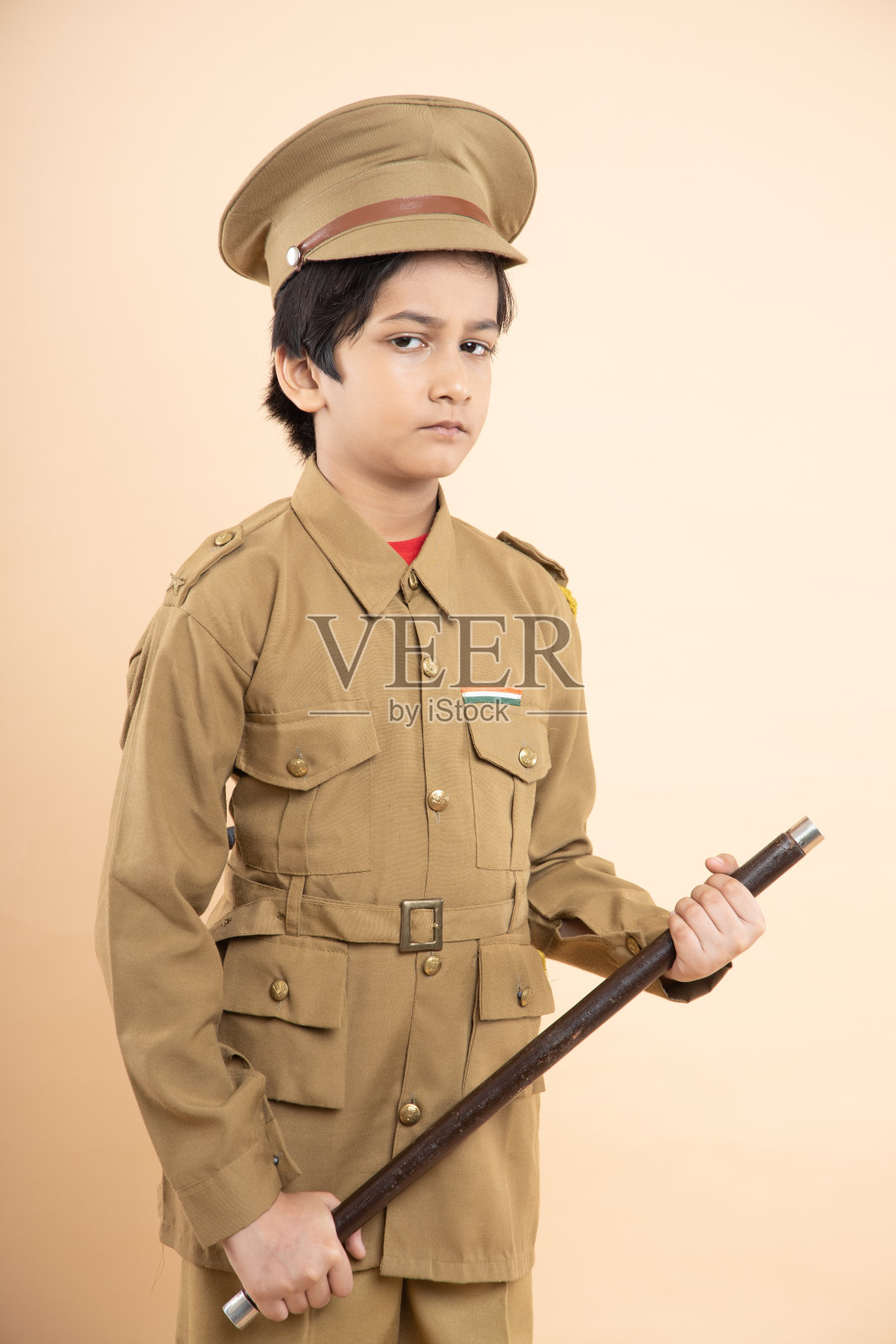 印度男孩穿着警服，手里拿着警棍，孤立在米色的工作室背景中。照片摄影图片