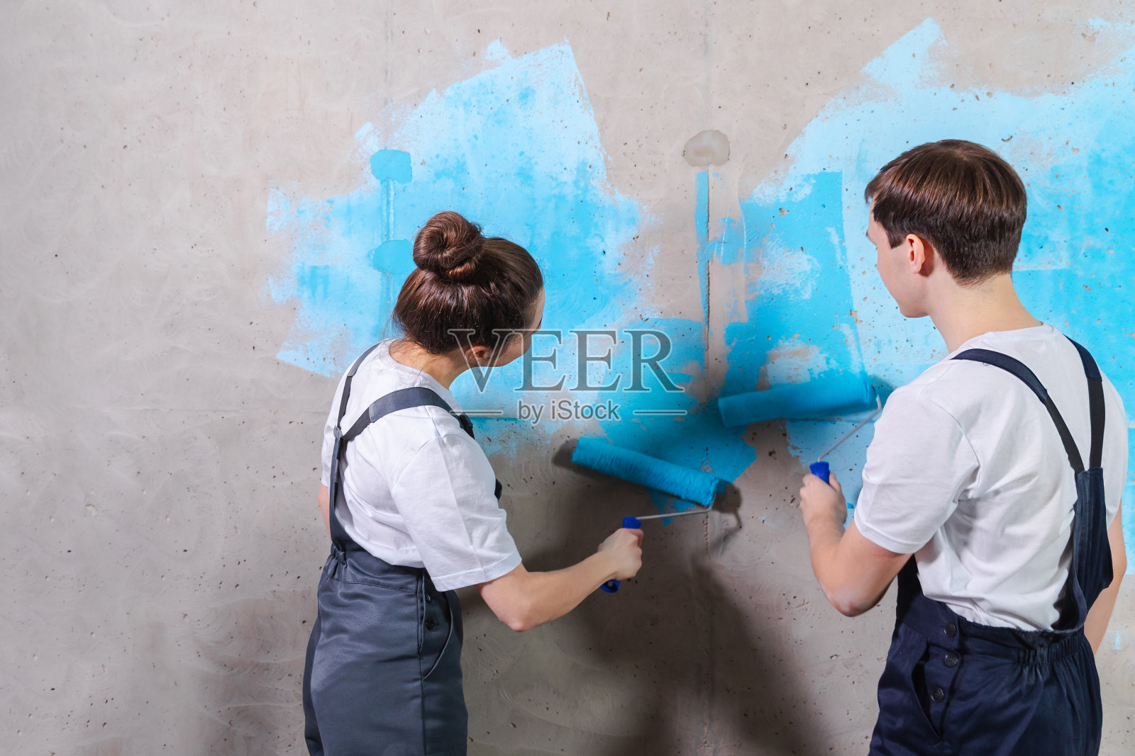 家居装修DIY装修理念。幸福的夫妇在新家的空房间里一起刷墙。丈夫和妻子在新房子里拿着油漆滚筒用蓝色油漆刷墙。照片摄影图片