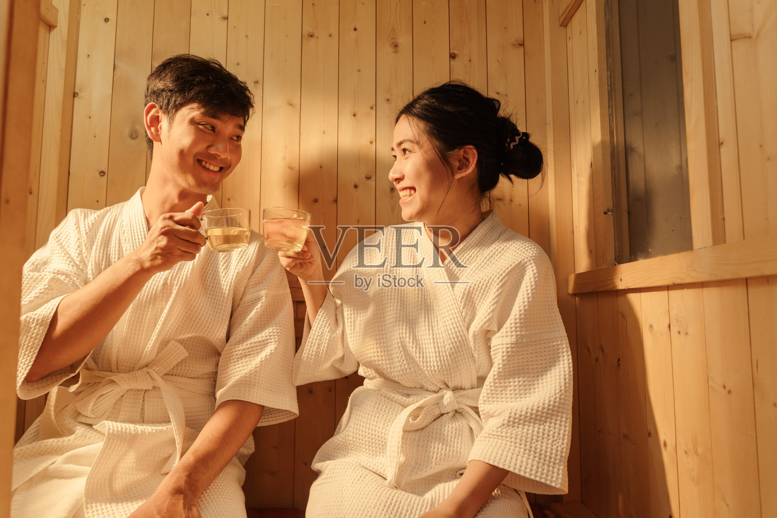 放松的亚洲夫妇穿着浴袍，一边喝着热茶，一边愉快地交谈，一边在桑拿浴室放松。健康和水疗治疗理念。照片摄影图片