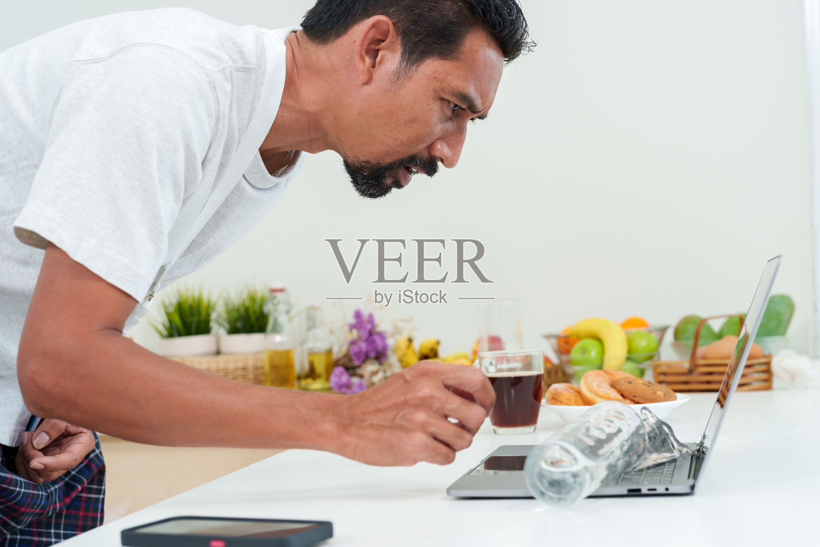 亚洲男人有胡子。一名男子将满满一杯水泼在笔记本电脑上，假装很震惊。在一天开始前喝咖啡的时候照片摄影图片