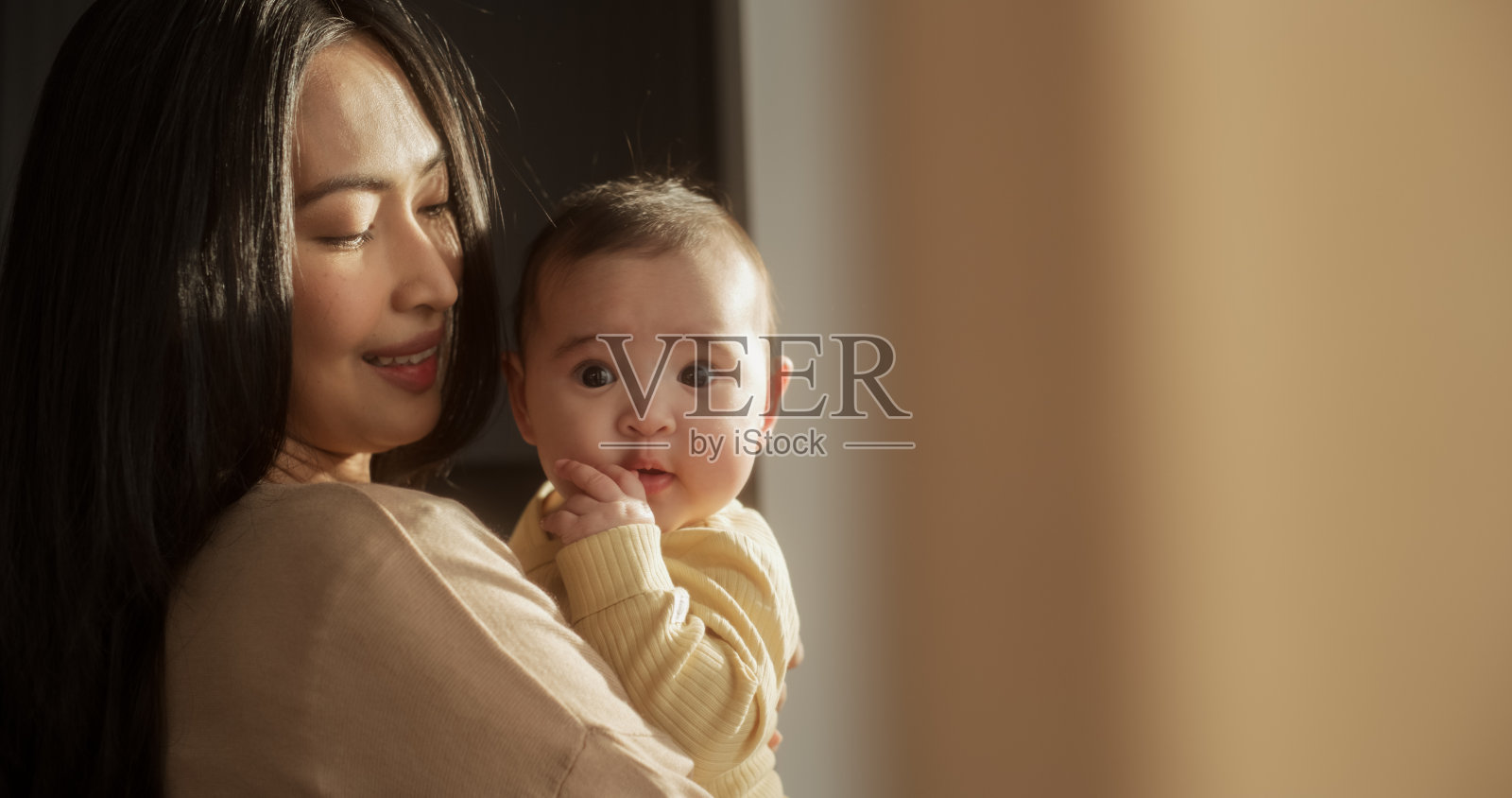 一个美丽的年轻亚洲妇女抱着她的孩子站在家里的窗户旁边的肖像。可爱的小男孩躺在妈妈的怀抱里，看着镜头照片摄影图片