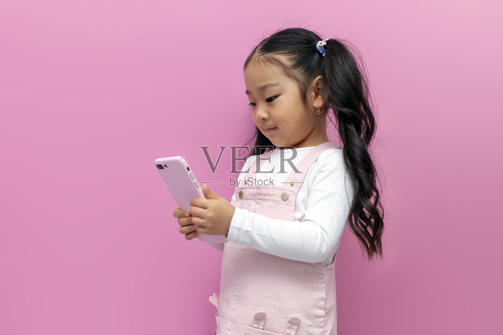 穿着粉色背心裙的亚洲小女孩使用智能手机，在粉红色孤立的背景下在线选择照片摄影图片