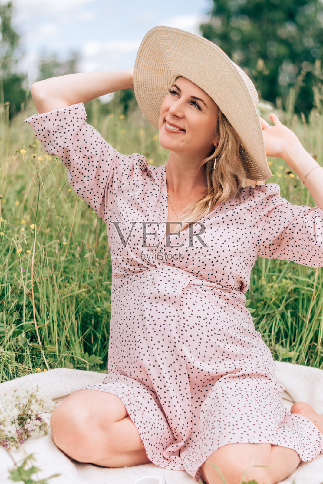 一个快乐的年轻金发孕妇穿着粉红色的裙子，戴着草帽，坐在一个夏天的草地上。夏季、孕期及产前护理理念照片摄影图片