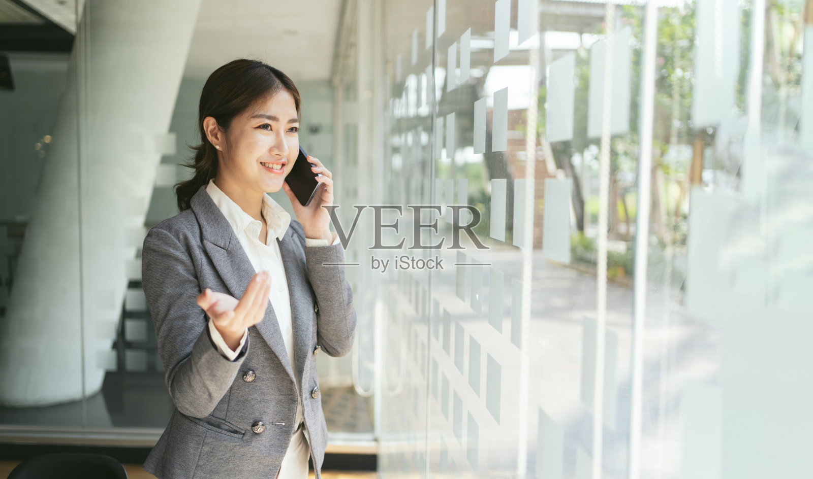 亚洲女性与智能手机站在现代办公背景和复印空间，时尚商业照片美丽的女孩在休闲西装与智能手机。照片摄影图片