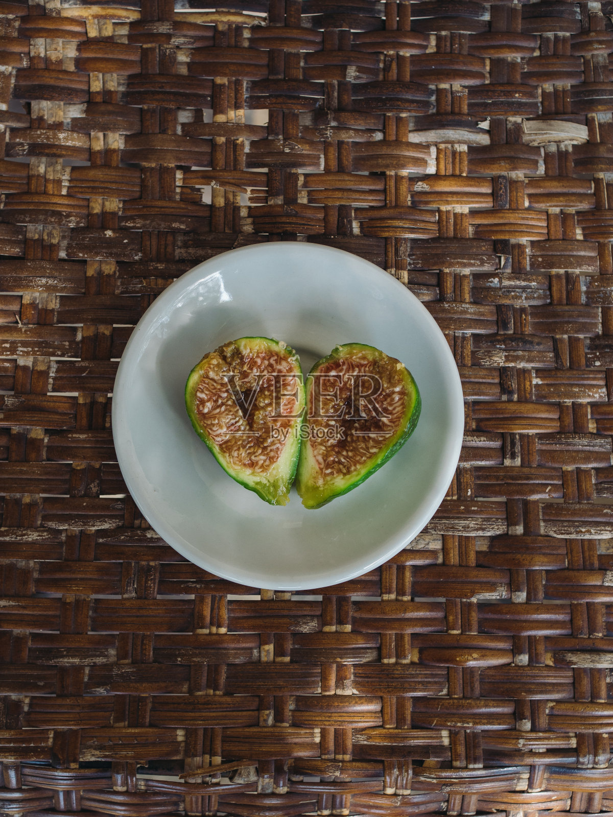 从上面垂直拍摄的两半成熟的绿色无花果，放在棕色稻草椅子上的白色盘子上。盛夏撒丁岛阳台的细节图。照片摄影图片