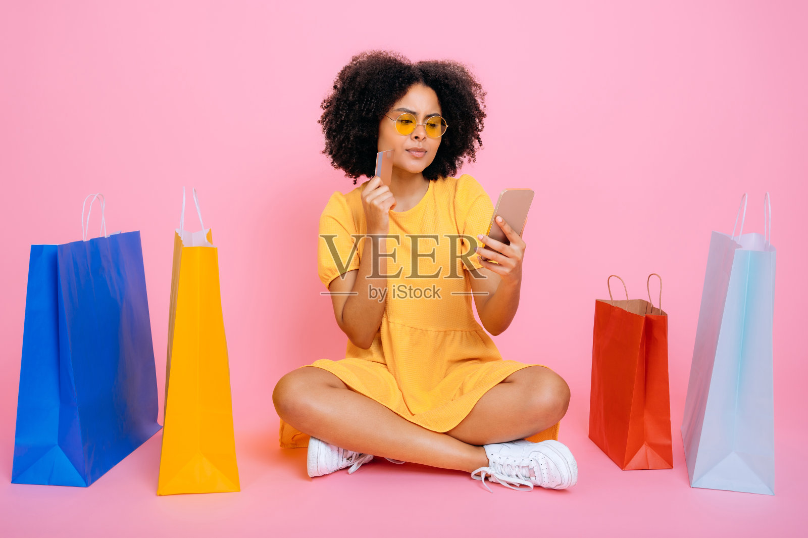 有思想的漂亮的混血卷发女人，穿着黄色的夏装，坐在装着购物的纸袋之间，拿着智能手机和信用卡，沉思地看着屏幕，孤立的粉红色背景照片摄影图片