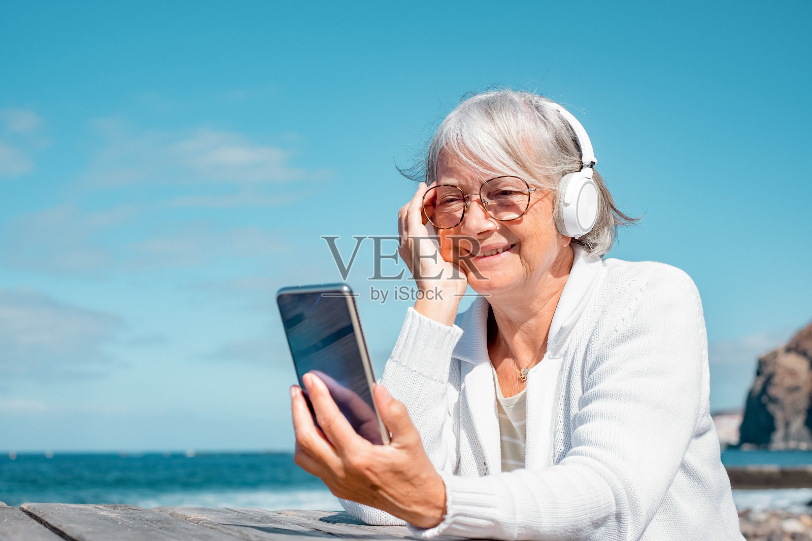 微笑的老妇人戴着耳机坐在海滩的木桌上，用手机享受科技和社交，大海和蓝天的地平线照片摄影图片