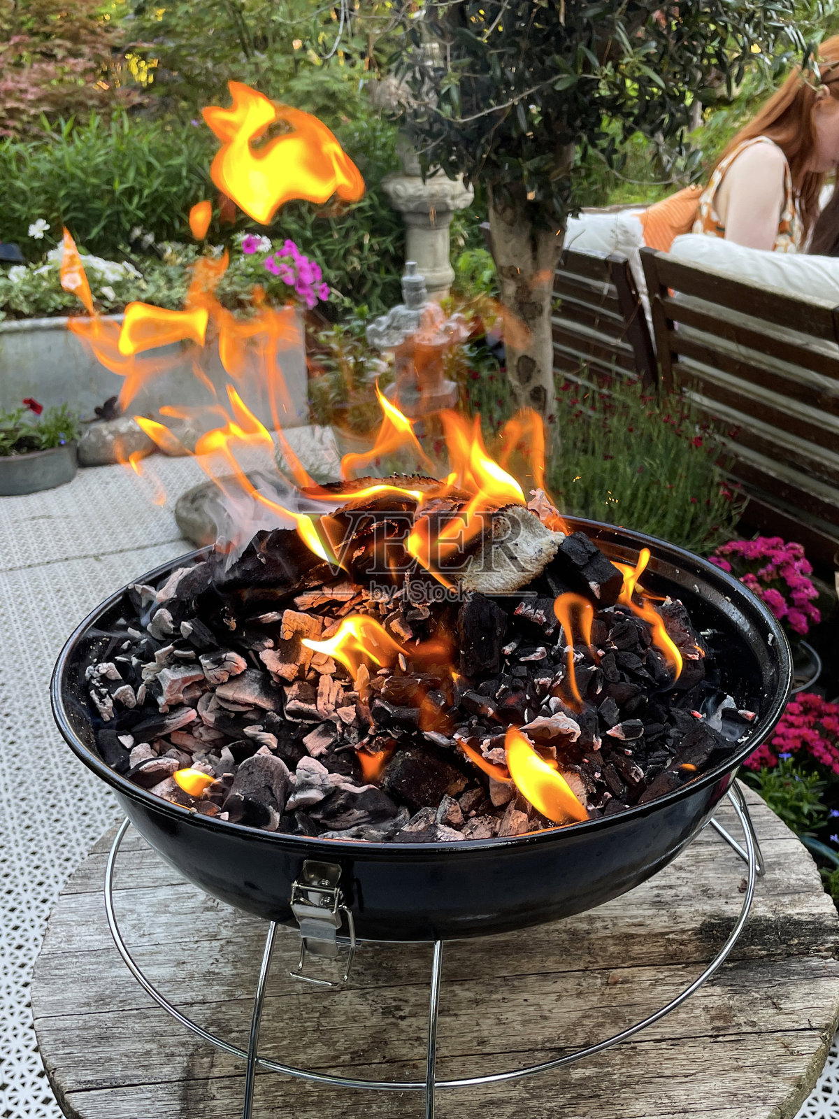 便携设备上燃烧的煤的特写图像，水壶，后花园的木炭烧烤，一群在户外休息区无法辨认的人，带靠垫的硬木座椅，盆栽开花植物，重点在前景照片摄影图片