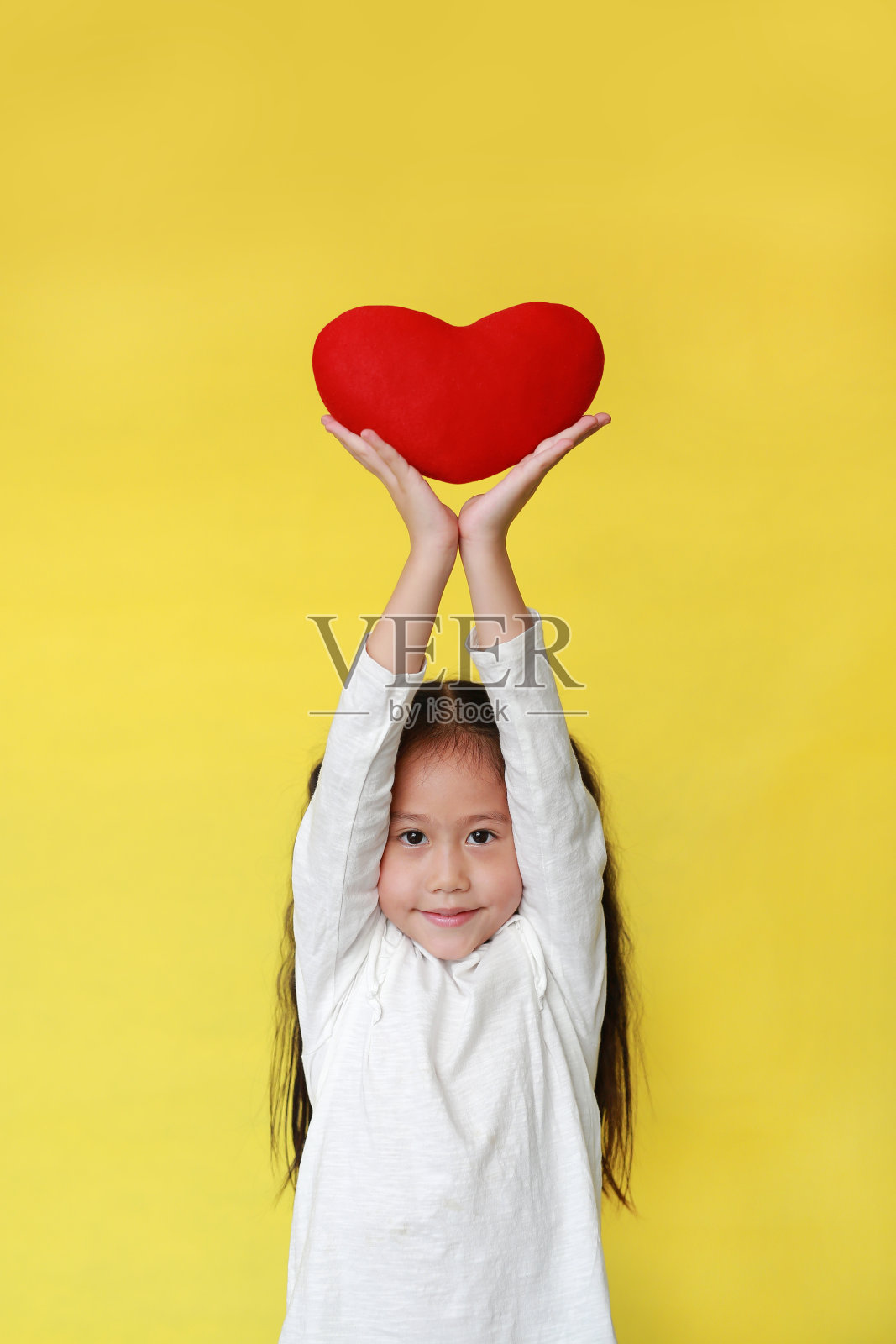可爱的亚洲小女孩举起红色的心在她的头上孤立的黄色背景。照片摄影图片