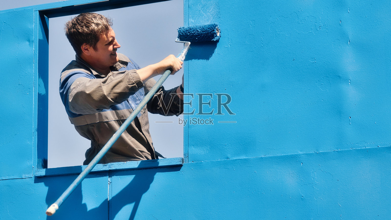 夏日里，穿着工作服的专业油漆工用油漆滚筒给建筑物的金属墙面刷漆。金属表面喷漆照片摄影图片