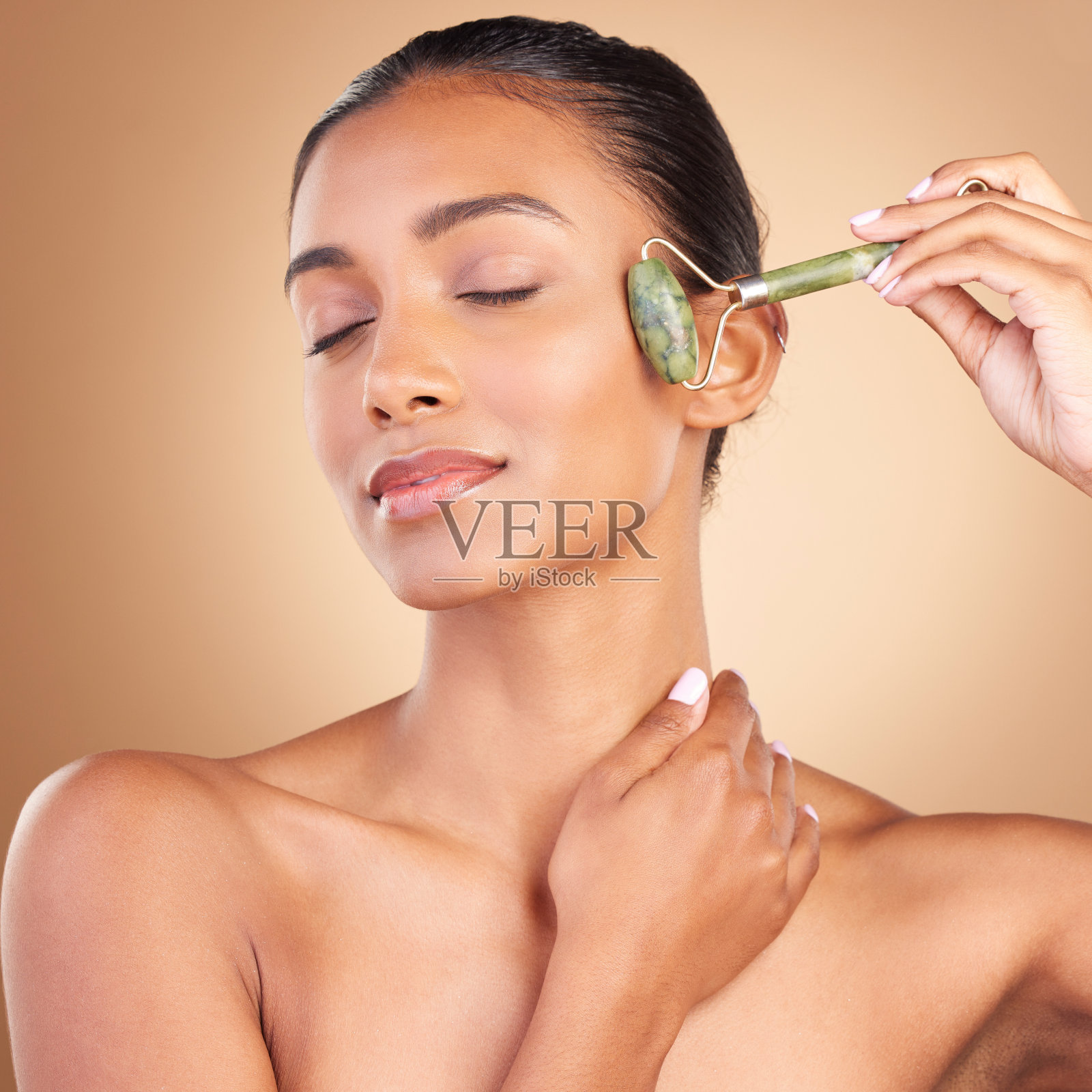 在棕色背景下，与模特女士在工作室里进行面部按摩和美容，自然抗衰老护肤。健康，奢华和产品与一个迷人的年轻女性使用滚轴皮肤提升照片摄影图片