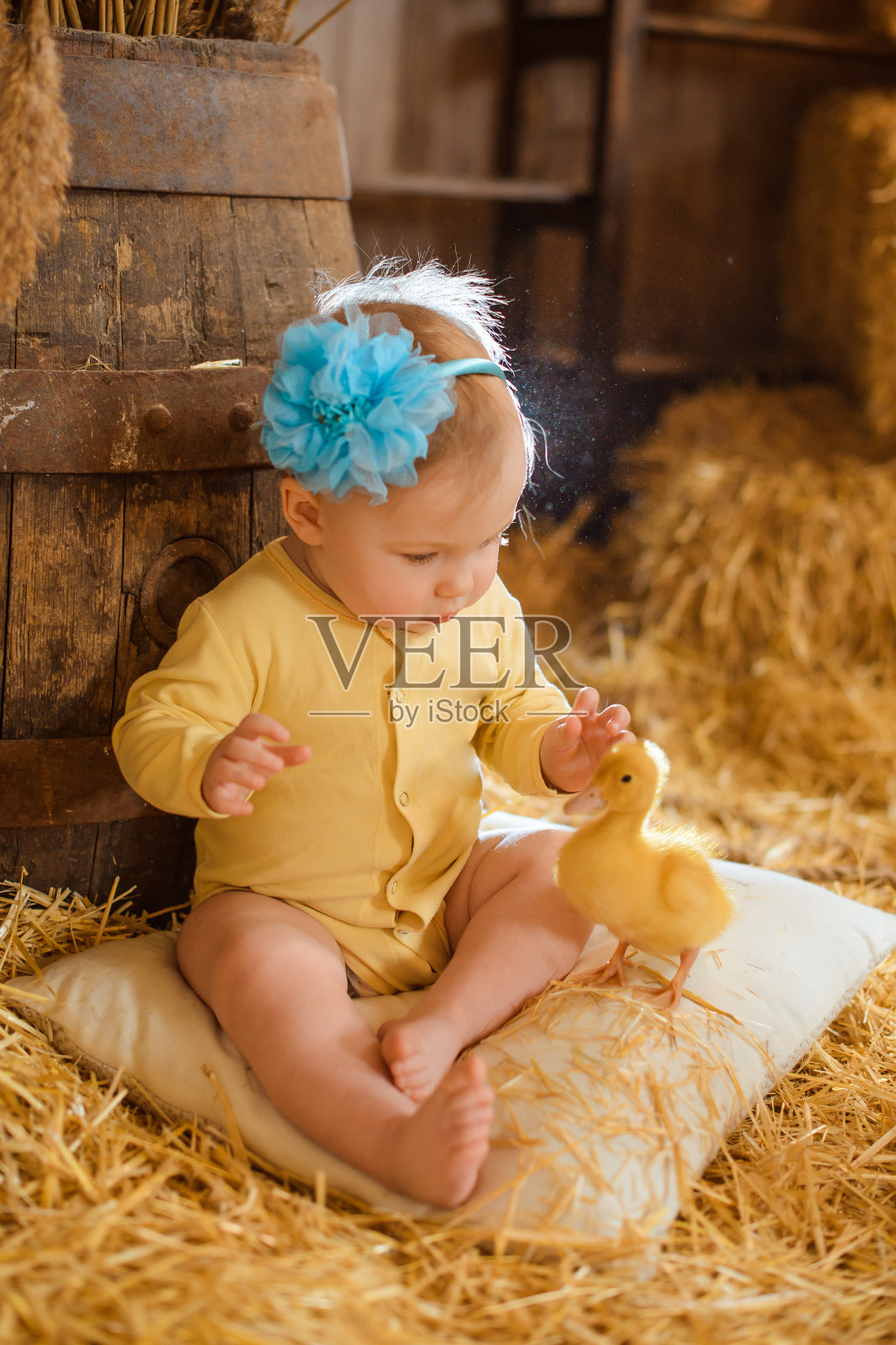 一个头上戴着蓝箍的小女孩坐在一个白色的枕头上，看着一只孤独的小鸭子，小鸭子也坐在枕头上，小女孩的身后是一个大桶。照片摄影图片