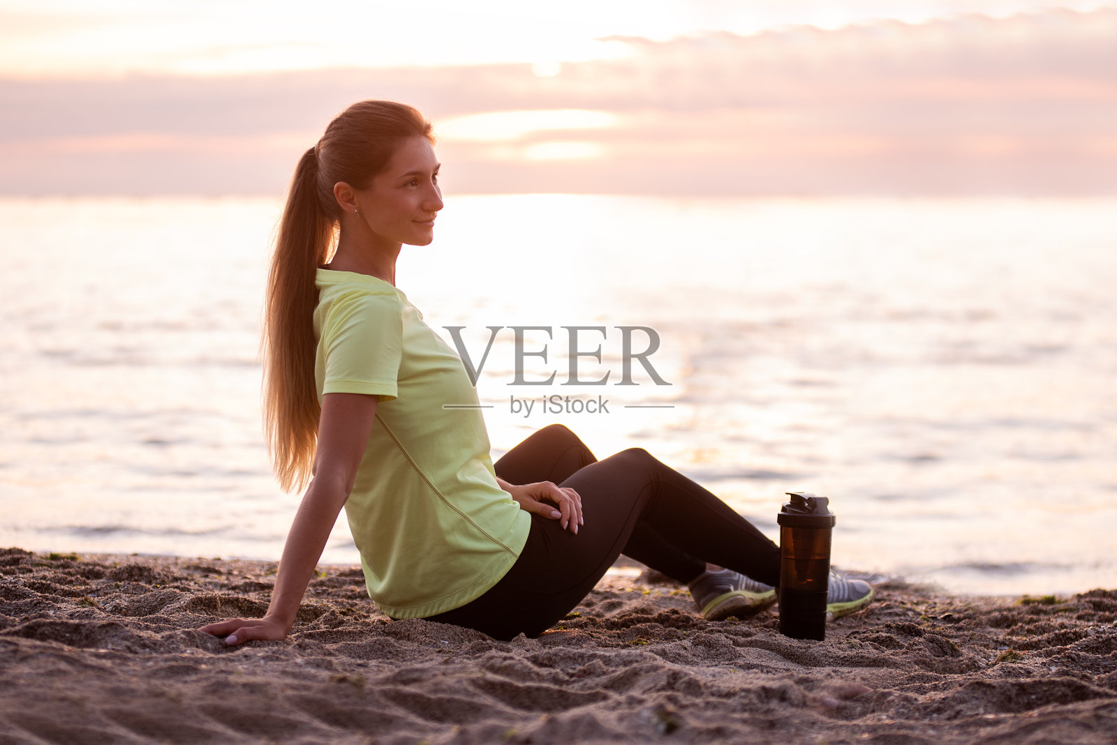日出时，穿着运动服的年轻女子坐在海边。女孩在锻炼后休息照片摄影图片