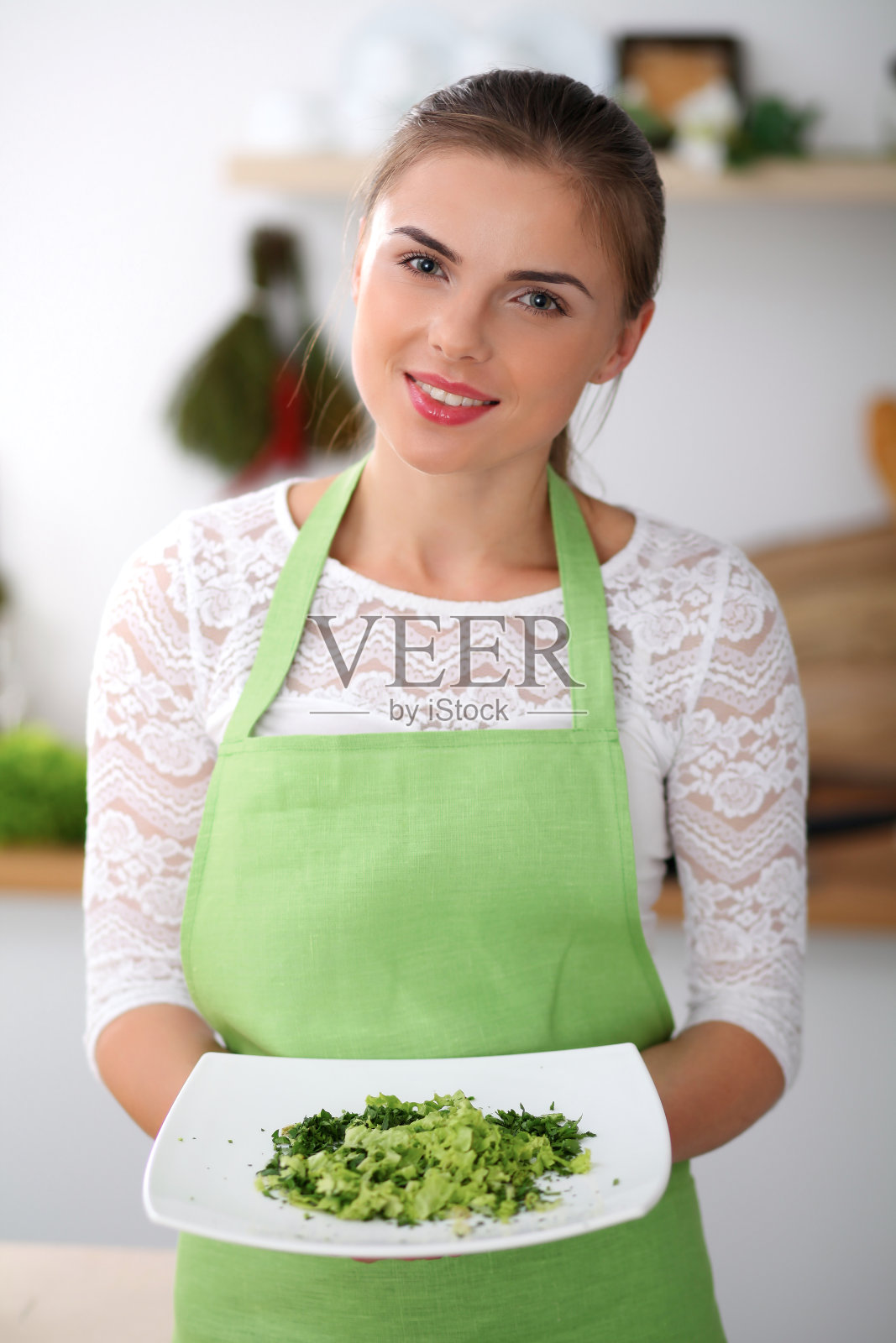 穿着绿围裙的年轻女子正在厨房里做饭。家庭主妇正在提供新鲜的沙拉。照片摄影图片