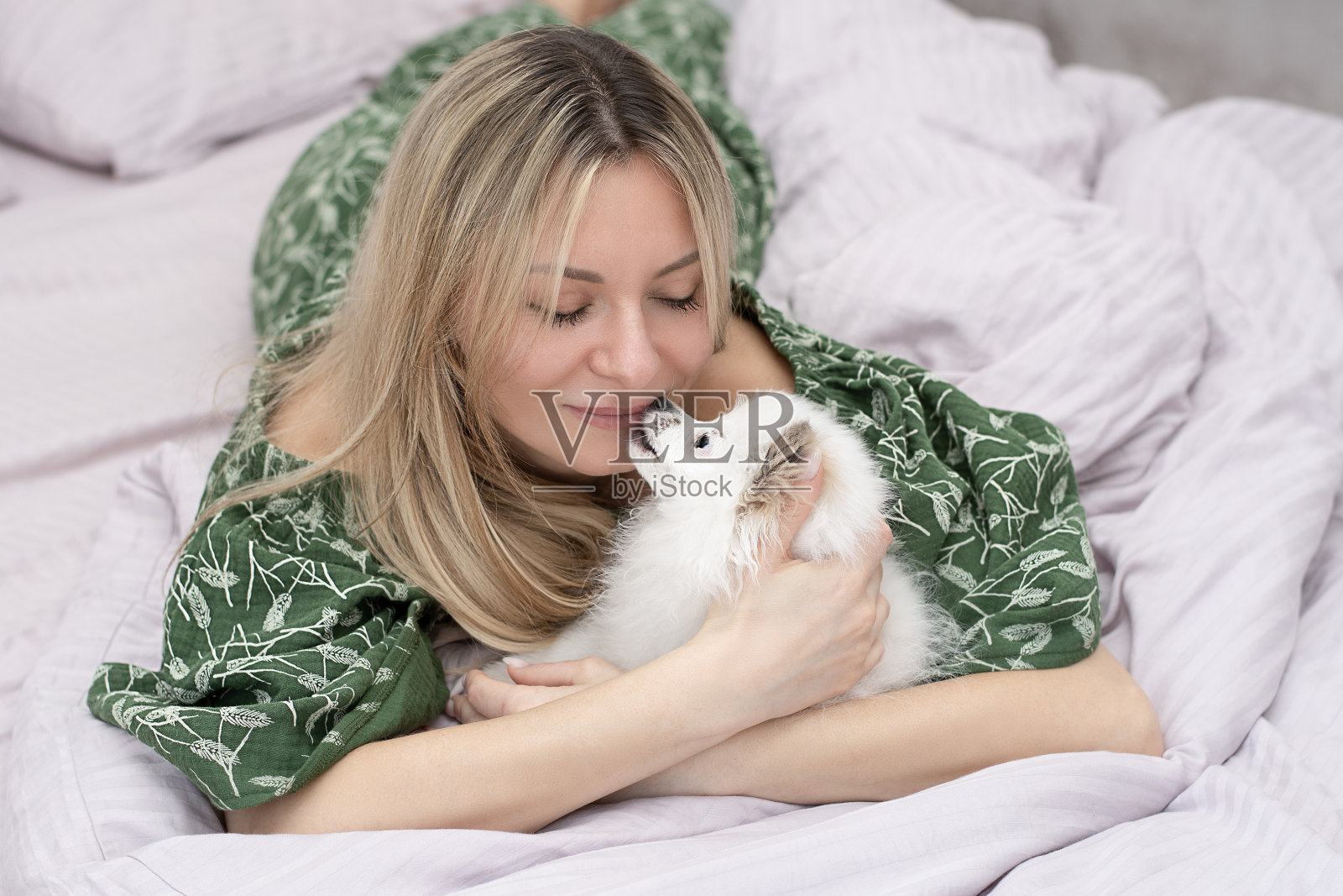 一位漂亮的年轻女子躺在家里的床上和一只毛茸茸的白色小猎犬玩耍。的概念。照片摄影图片