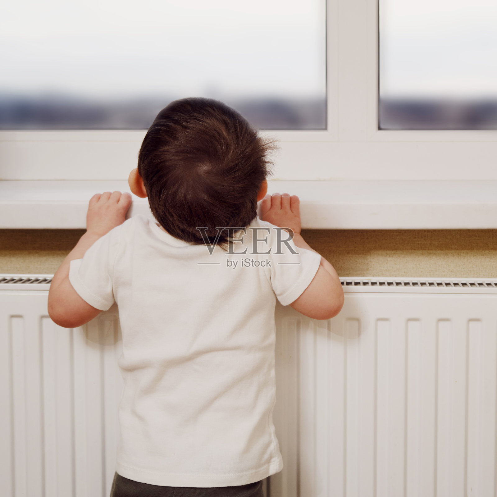 蹒跚学步的婴儿爬到靠近加热电池的窗户上。一个小孩试图爬上窗台。一岁八个月的孩子照片摄影图片