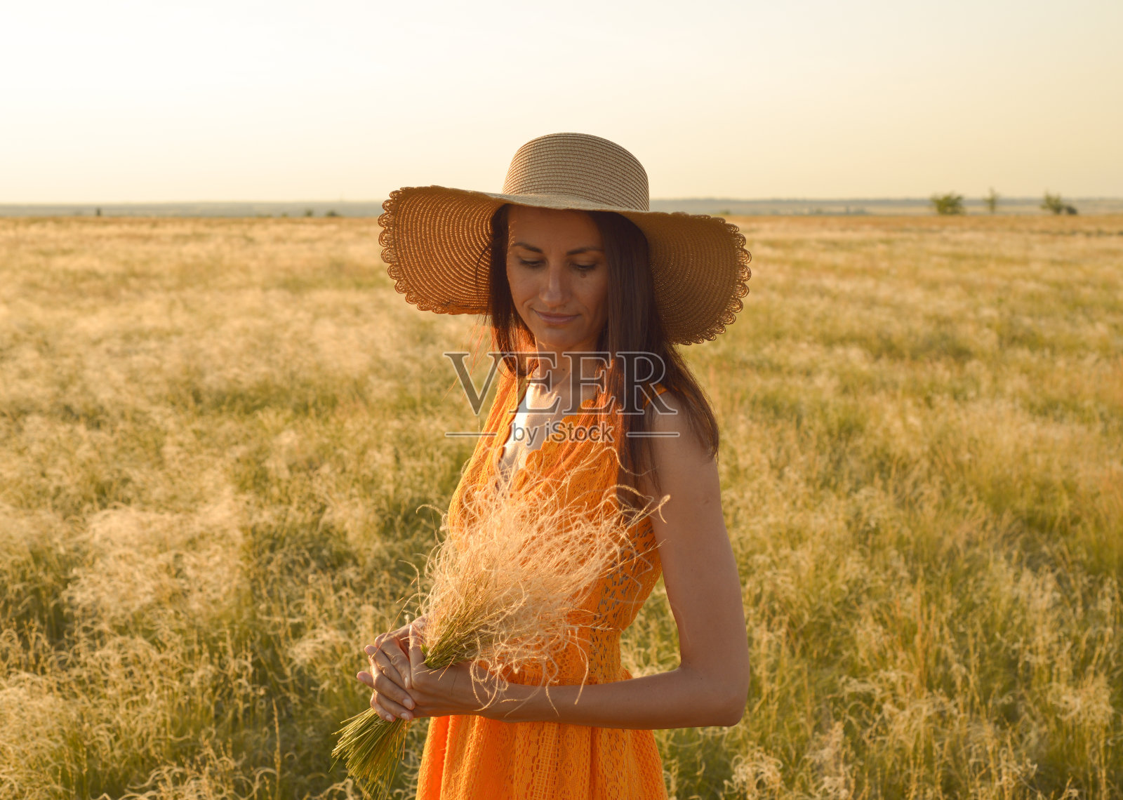 身穿橙色连衣裙，头戴草帽的年轻女子站在夕阳下的田野里照片摄影图片
