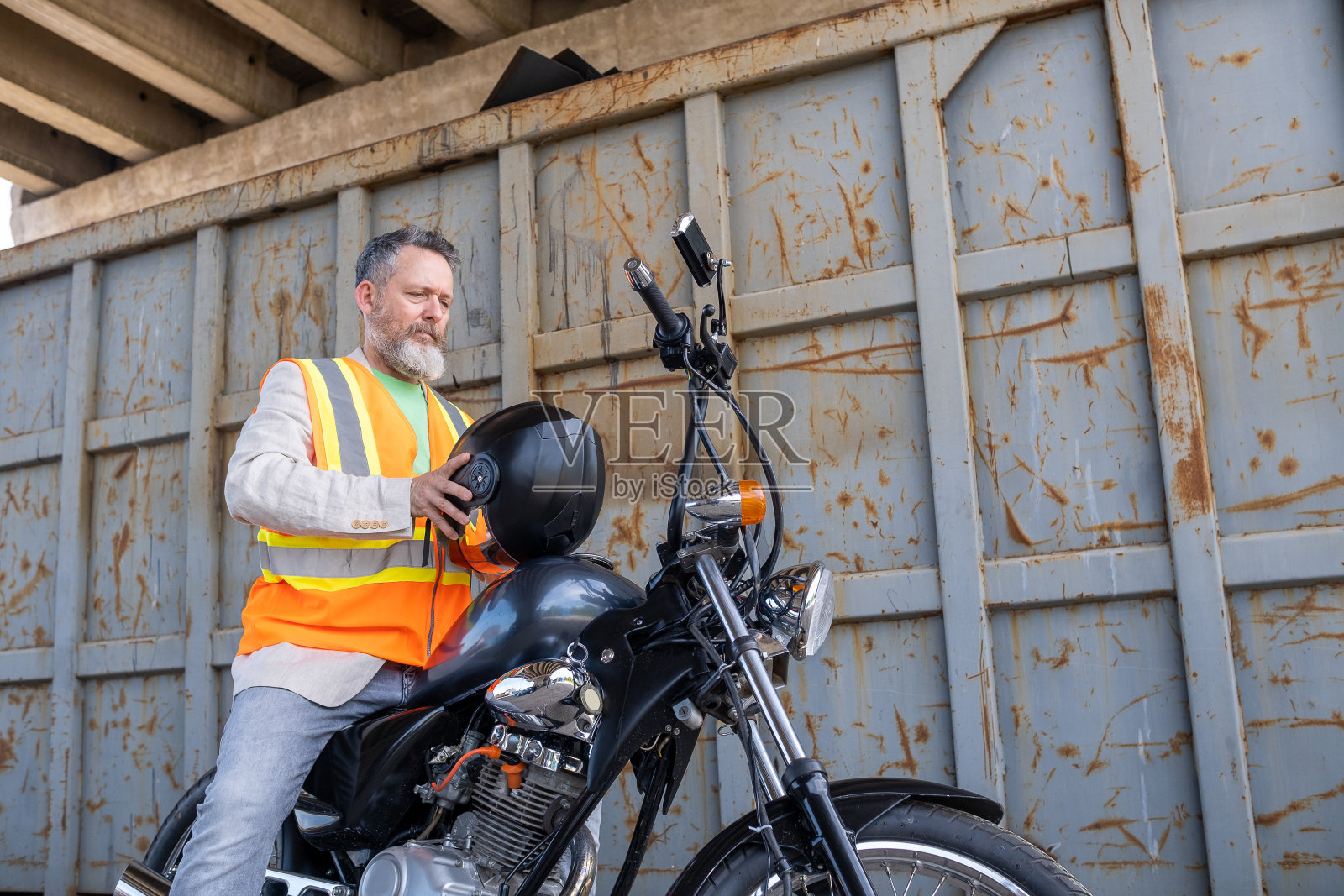 一个成熟的男人，穿着浅绿色的t恤，米色的夹克，灰色的牛仔裤和反光背心，骑着一辆摩托车。照片摄影图片