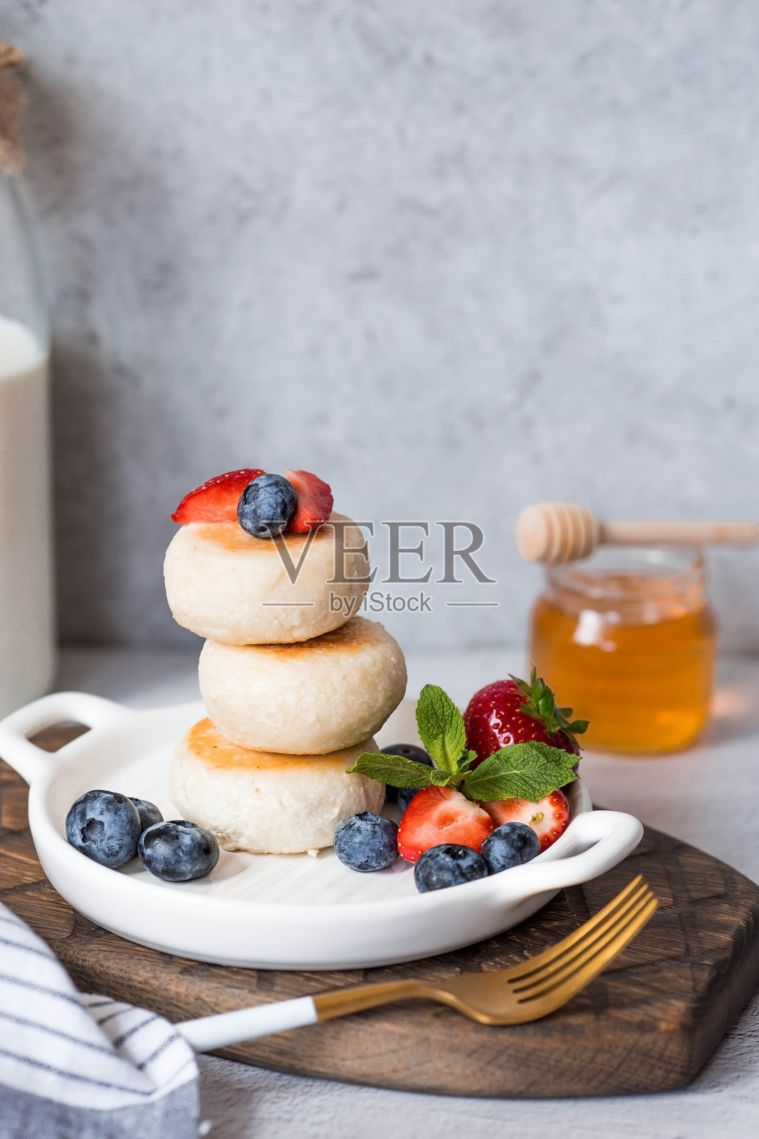 灰色混凝土桌子上时髦的木板上放着松软干酪煎饼、俄罗斯奶酪蛋糕和新鲜的蓝莓、草莓、蜂蜜和薄荷叶。自然产品。健康美味的早餐。照片摄影图片