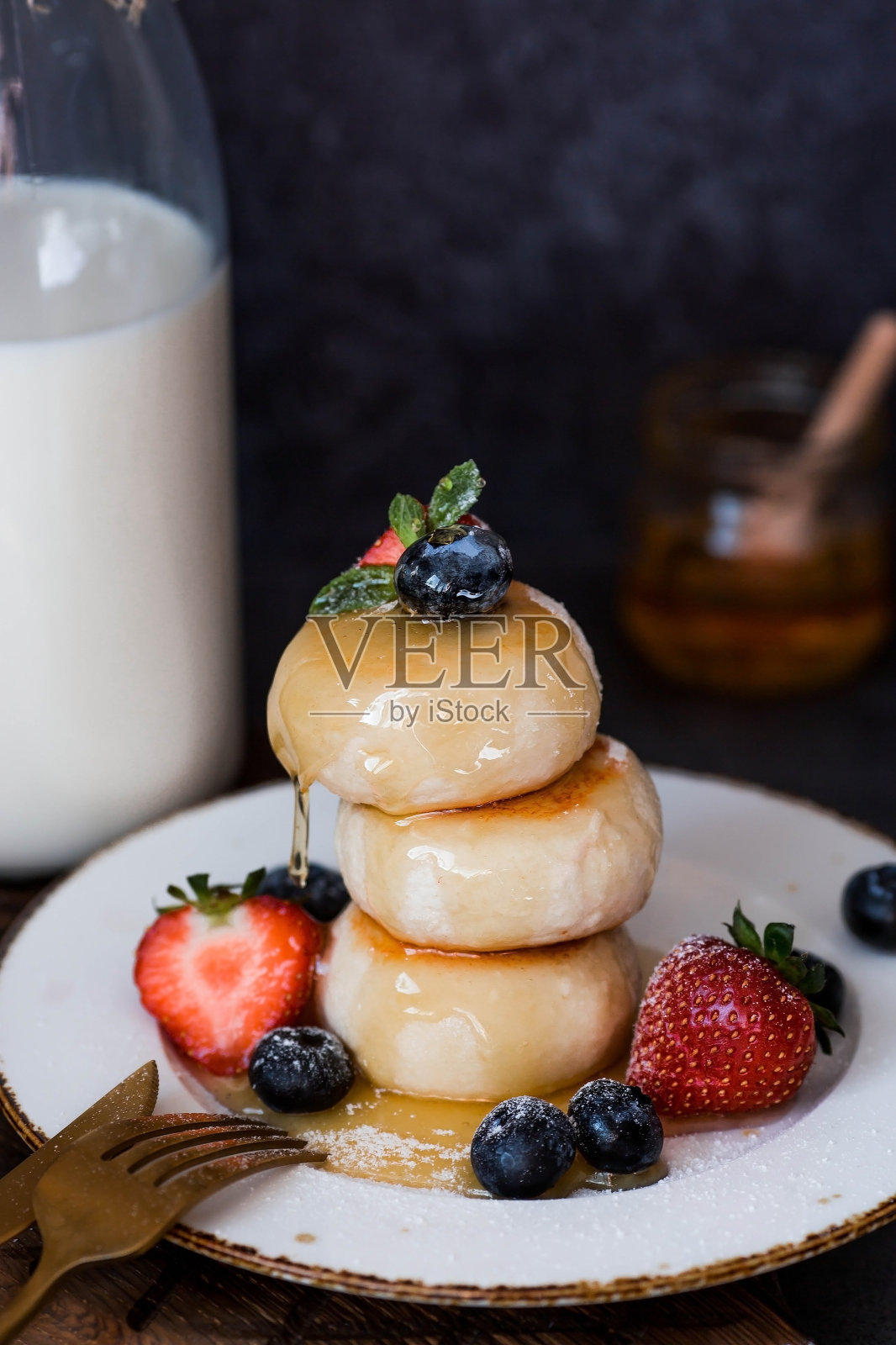 农舍奶酪煎饼，俄罗斯奶酪蛋糕，新鲜蓝莓，草莓，蜂蜜和薄荷叶放在一个时尚的深色背景的木板上。自然产品。健康美味的早餐。照片摄影图片