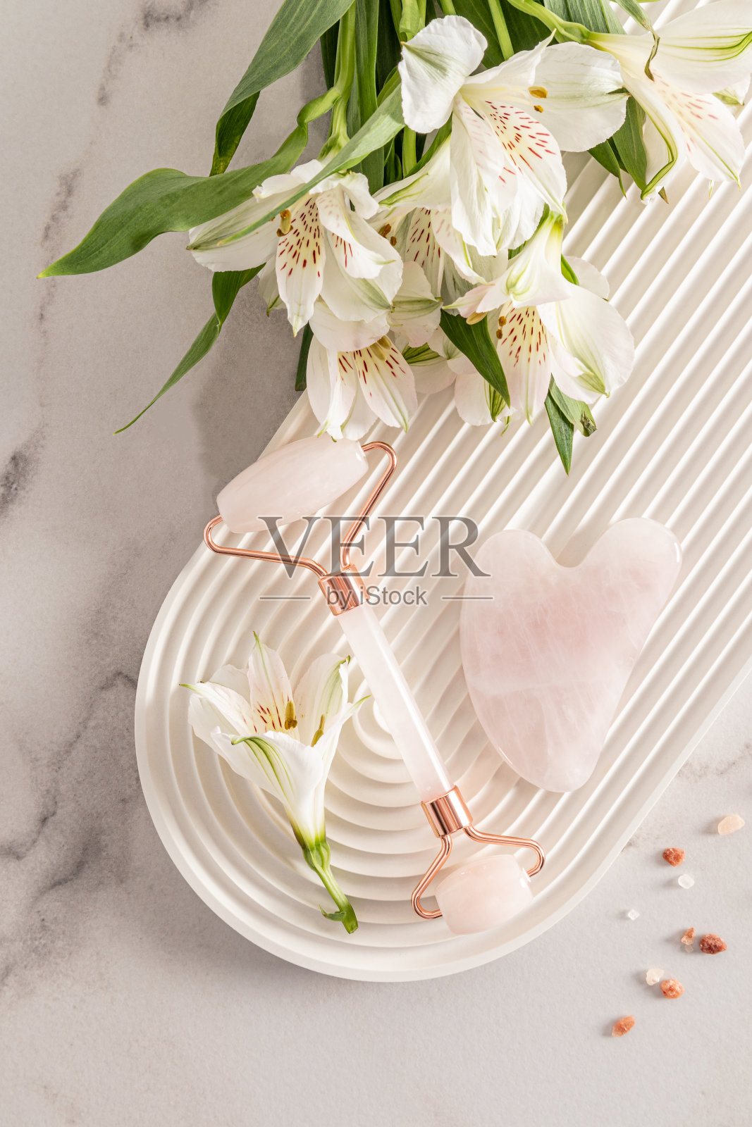 一个玫瑰石英化妆滚轮按摩器，一个瓜沙刮刀和天仙花躺在一个白色的拱形基座上。前视图。自我保健。照片摄影图片