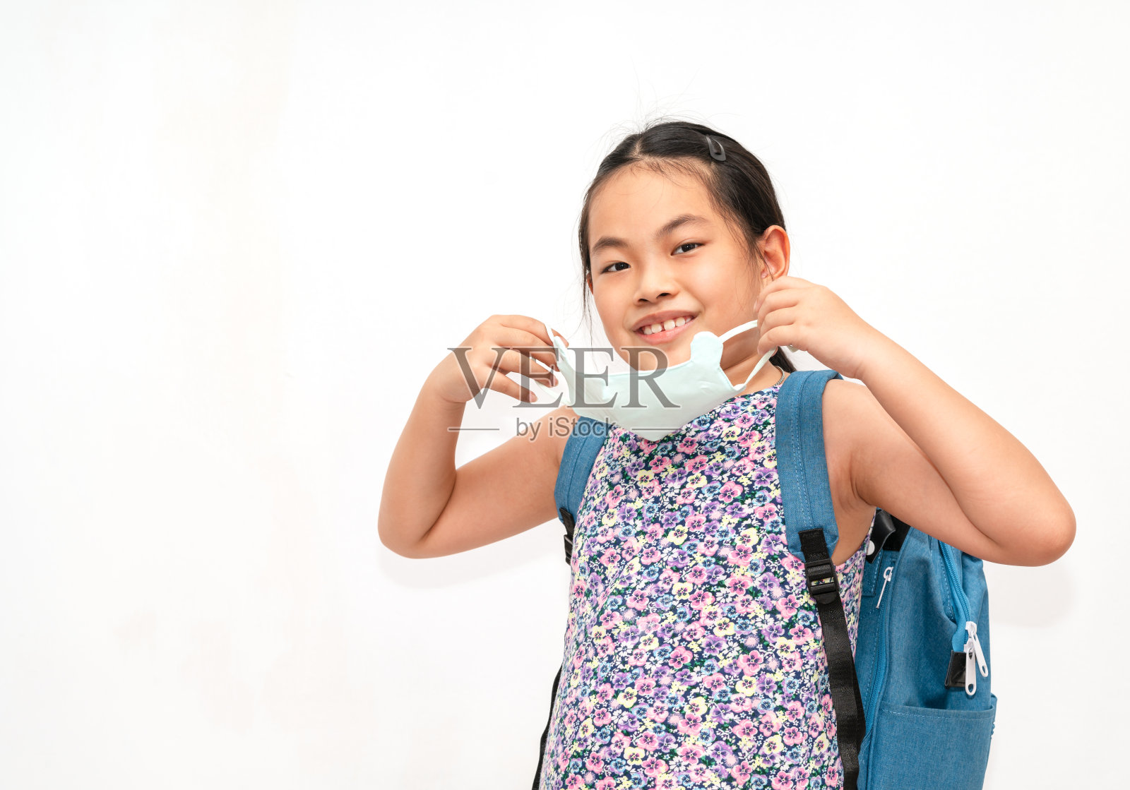 画像上的亚洲小女孩正抱着并愿意戴上口罩，背着双肩包去上学，孤立的白色背景肖像。返校的概念。照片摄影图片