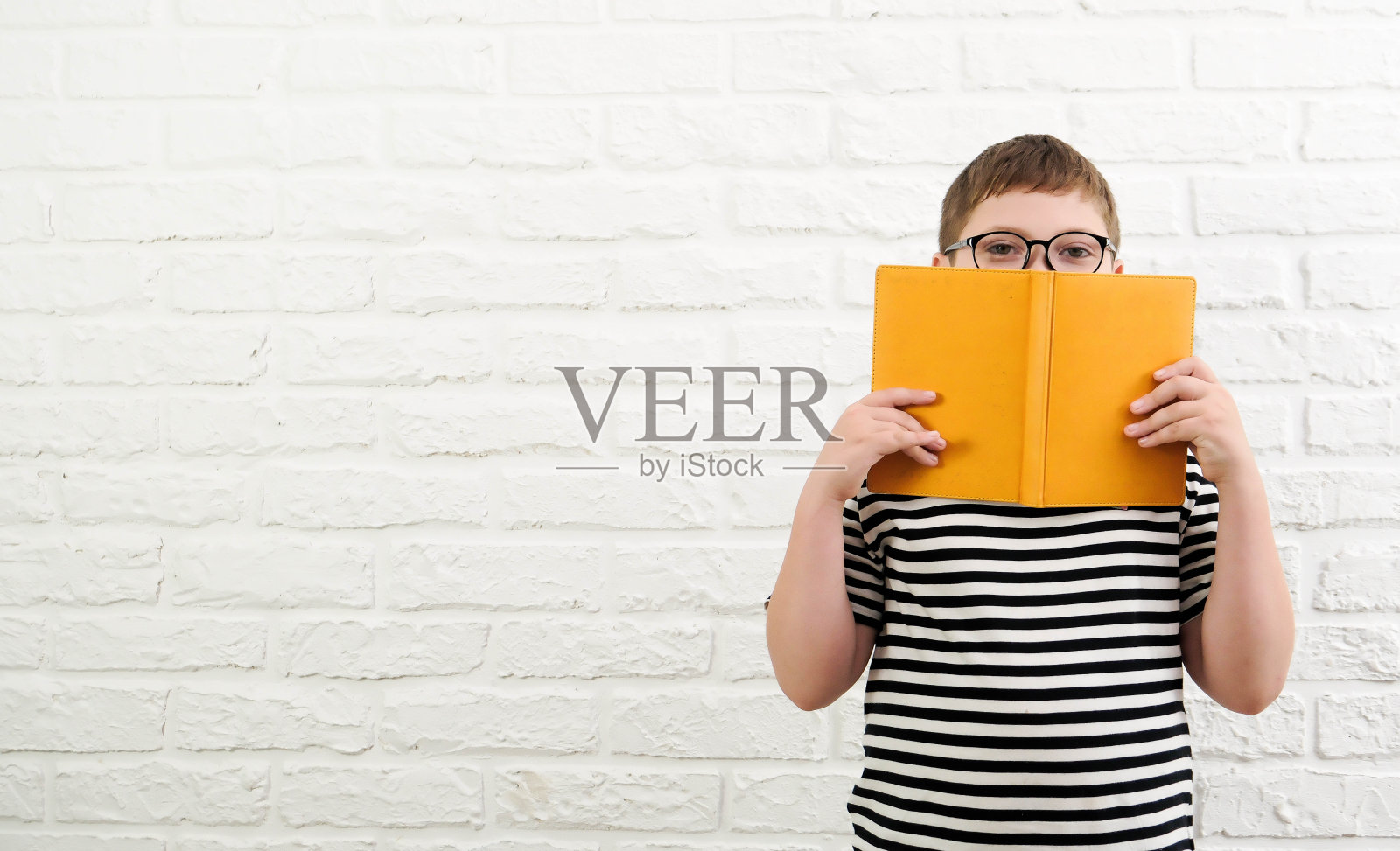 一个可爱开朗的男孩，穿着条纹t恤，戴着眼镜，拿着一本黄色的书，半脸贴在白砖背景上。照片摄影图片