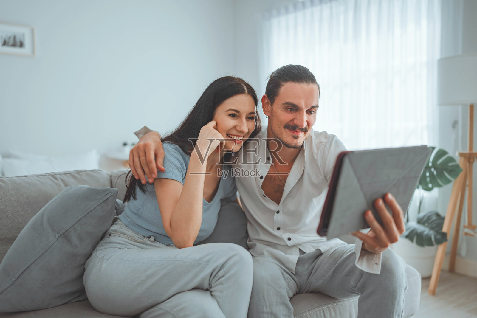 深情的年轻人抱着迷人的妻子女友，看着平板电脑屏幕，男男女女在舒适的沙发上放松。网上购物的情侣申请照片摄影图片