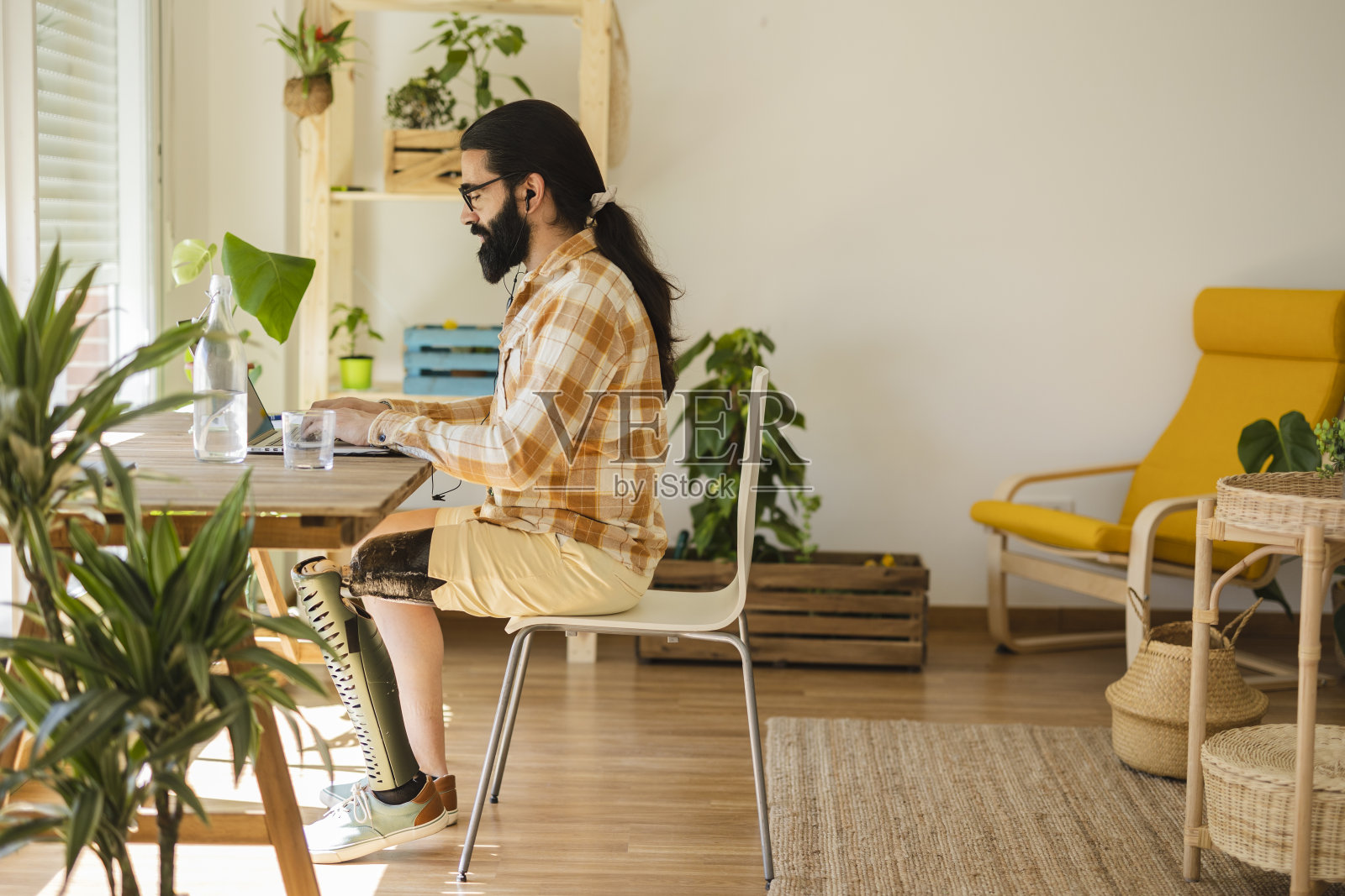 带着假腿的普通成年男子在家里办公桌旁使用笔记本电脑，远程工作。满脸胡须的快乐男人微笑的肖像。商人在家管理互联网业务照片摄影图片