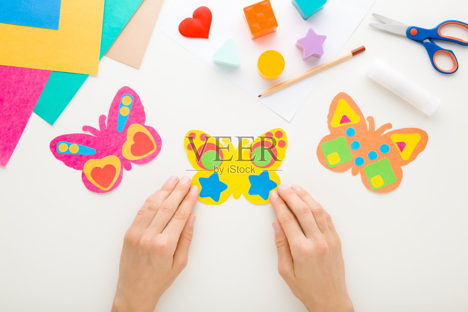 年轻的成年妇女手拿着彩色蝴蝶形状的纸在白色的桌子背景。柔和的颜色。制作装饰元素。特写镜头。视角镜头。自顶向下视图。照片摄影图片