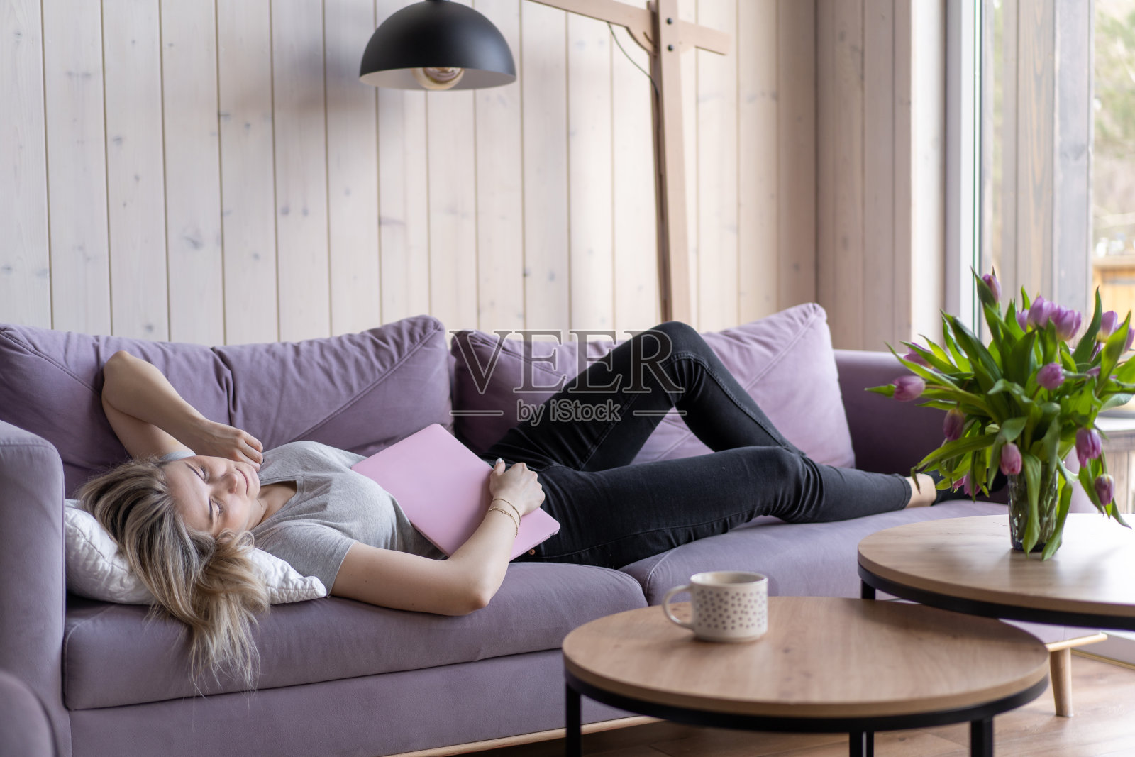 睡眠问题。穿着粉色t恤和牛仔裤的金发美女手里拿着笔记本电脑在家里的沙发上睡着了。在家上网工作。周末。过度劳累。乏力照片摄影图片