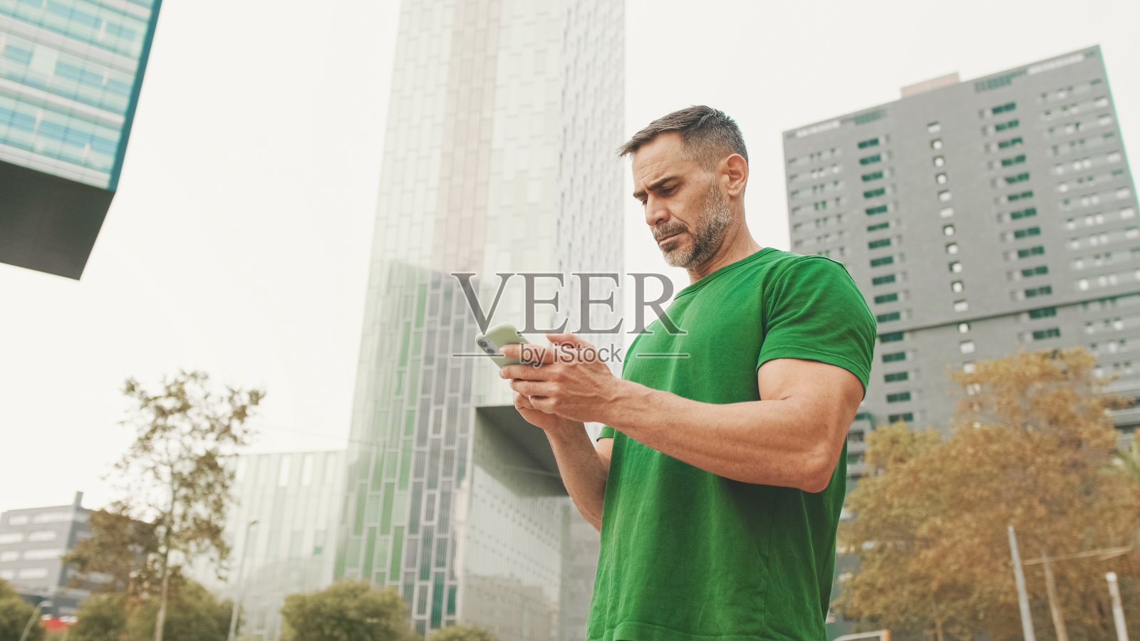 成熟的男性游客穿着休闲服，站在现代建筑的背景上，用智能手机寻找正确的方向，使用地图应用程序照片摄影图片