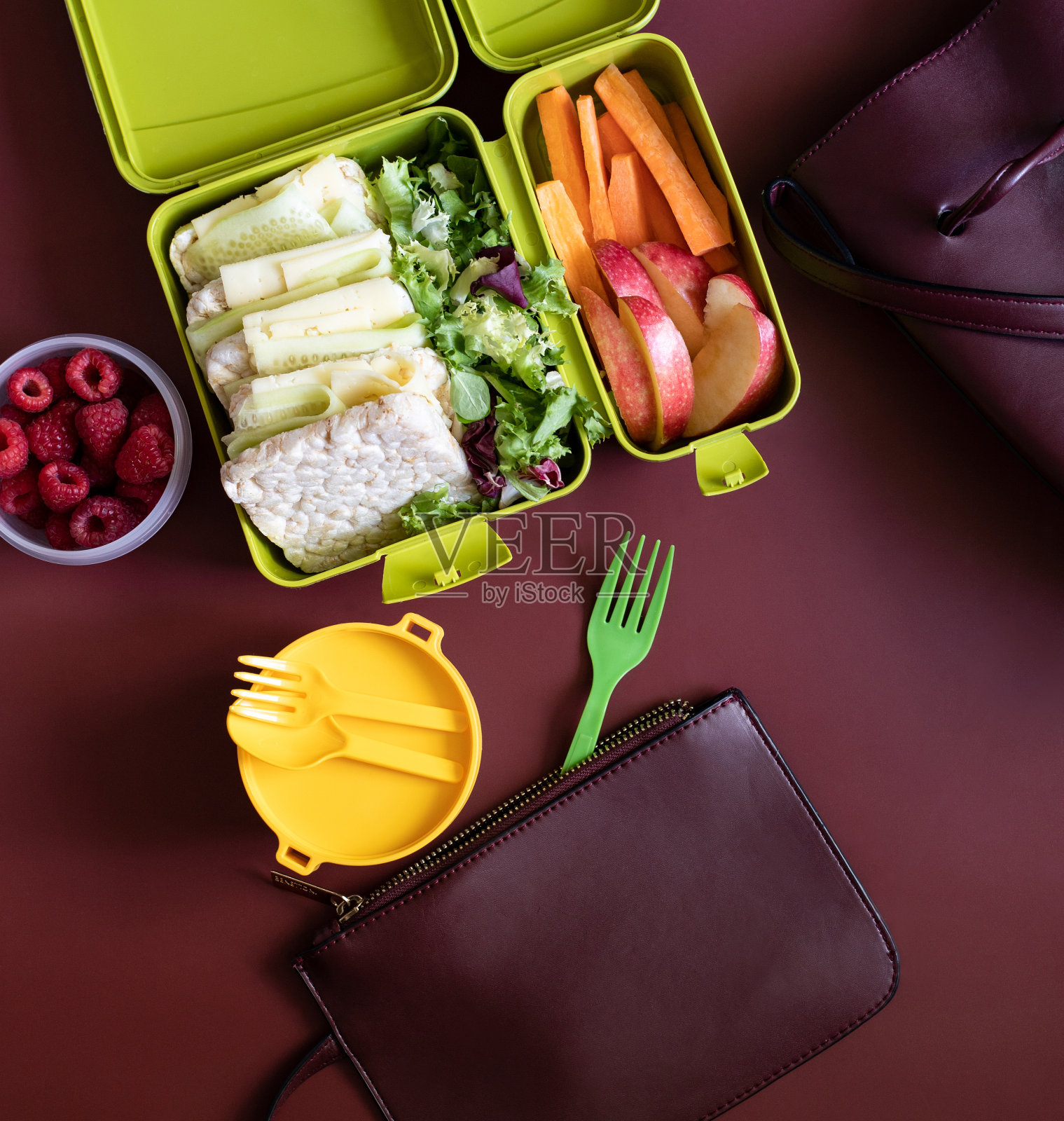 午餐盒里有三明治、胡萝卜条、苹果、生菜和覆盆子，以酒红色为背景，还有一个女人的手提包和钱包。健康食品的概念。俯视图照片摄影图片