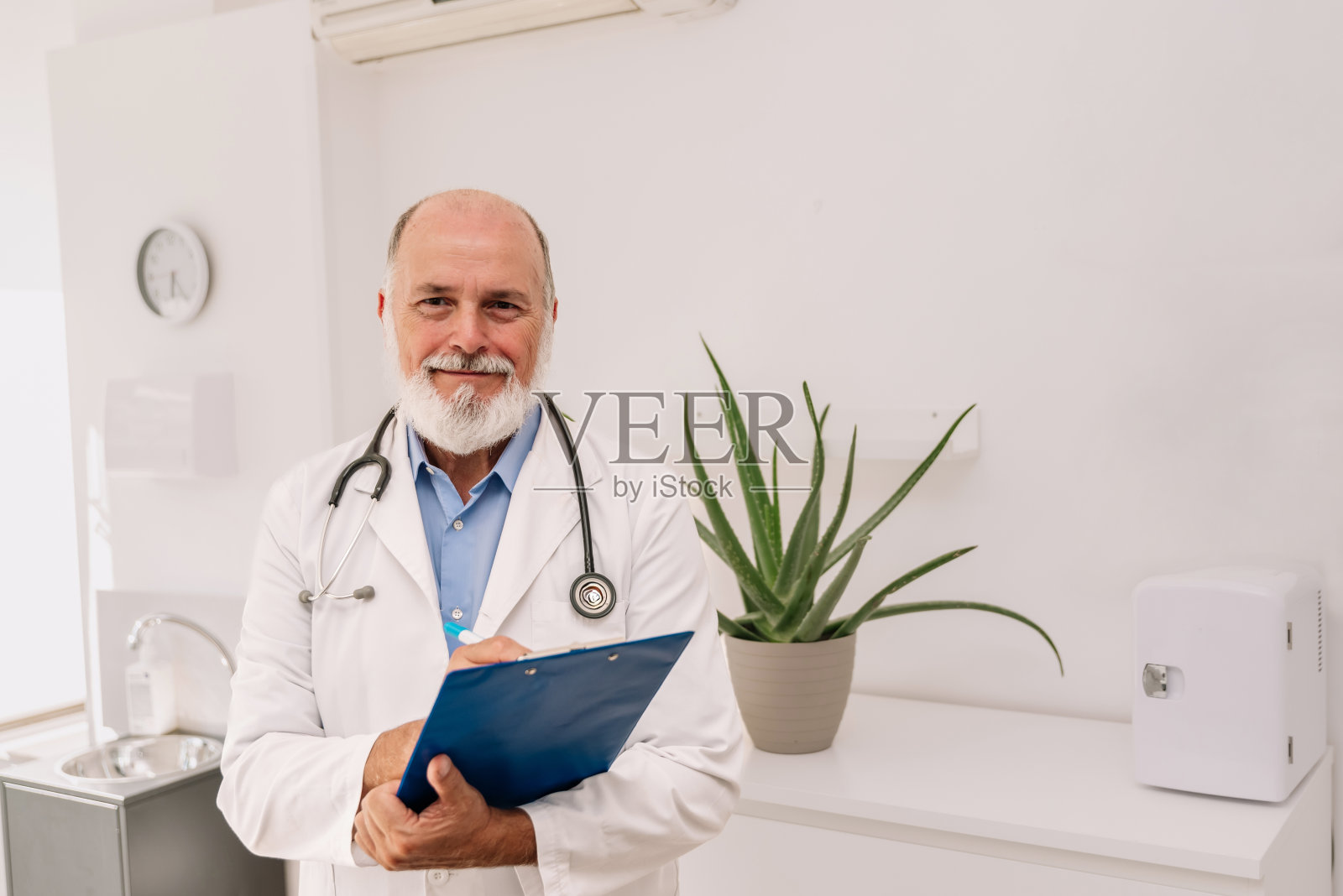 资深男医生，胡子灰白，双臂交叉，穿着长袍，在诊所里拿着听诊器看着摄像机照片摄影图片