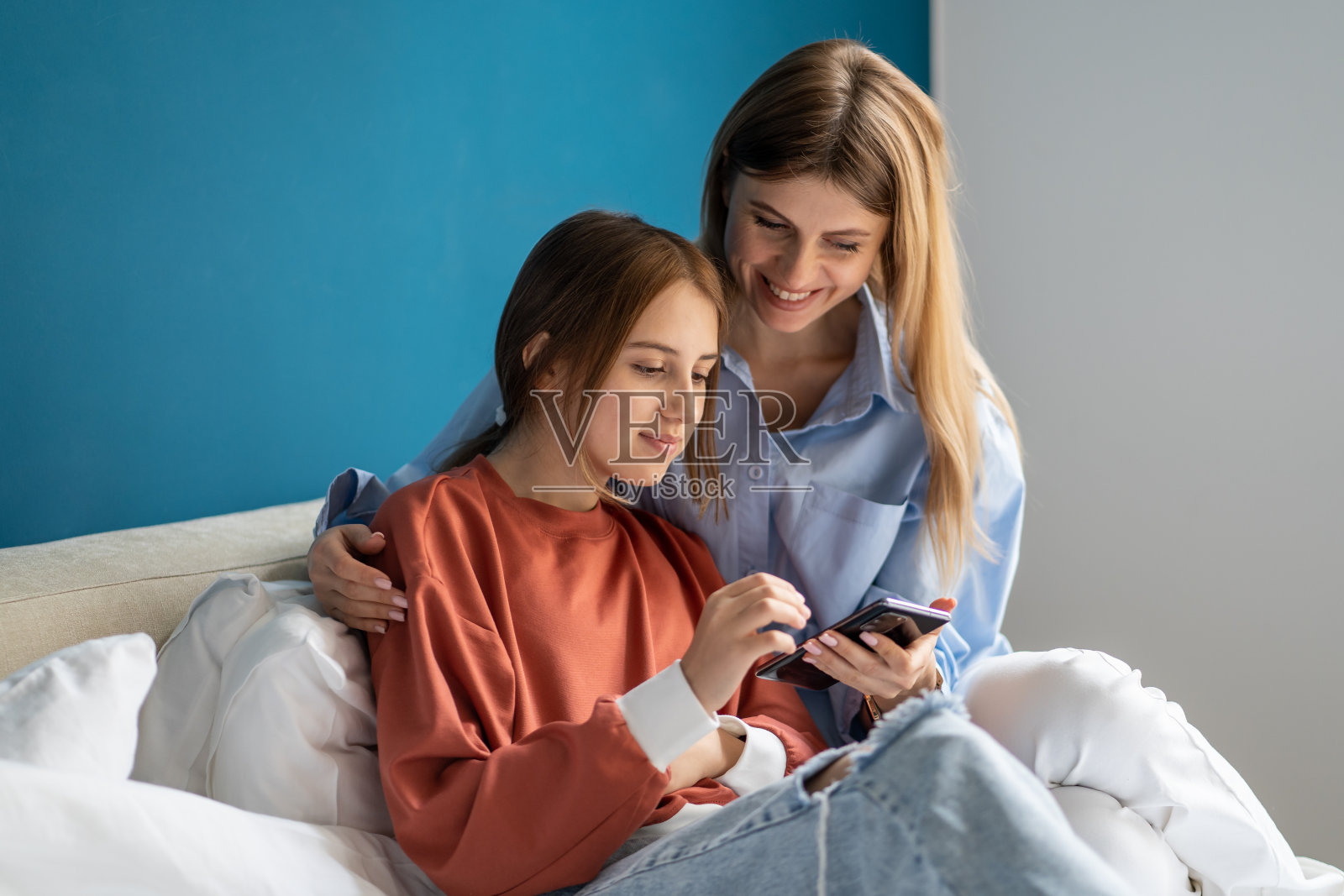 无忧无虑微笑的母亲和十几岁的女儿一起在床上放松地玩智能手机照片摄影图片