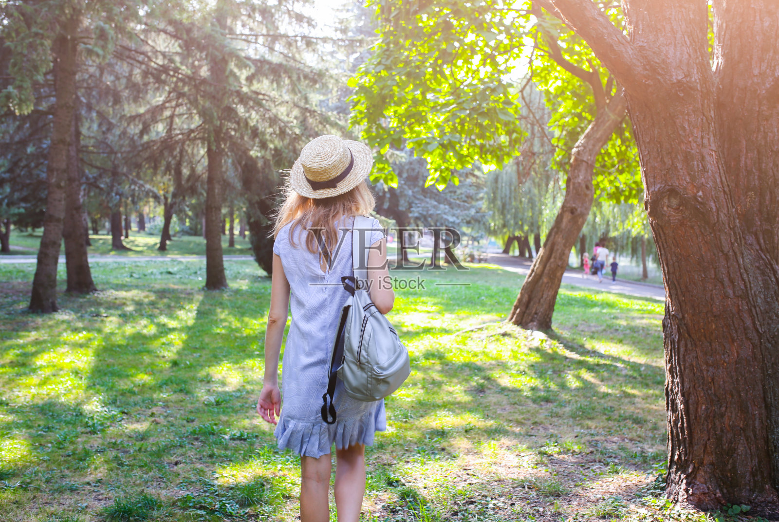 戴着草帽的时髦女孩站在户外。穿蓝裙子的女人。银灰色背包。旅游度假概念。夏日生活写真。阳光和树木的阴影。照片摄影图片
